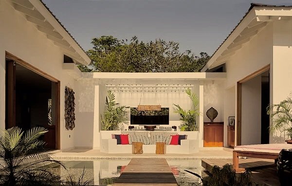 A modern outdoor patio with a fire pit, surrounded by white walls, tropical plants, and seating with red cushions, overlooking a pool.