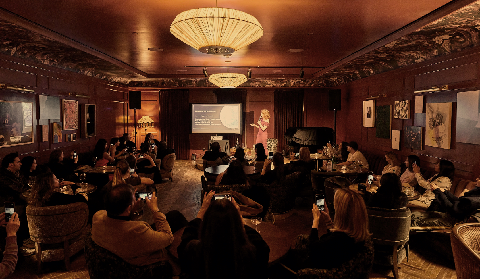A woman is giving a presentation to an audience in a dimly lit room with wooden walls, artwork, and large chandeliers. Some audience members are taking photos with their phones.