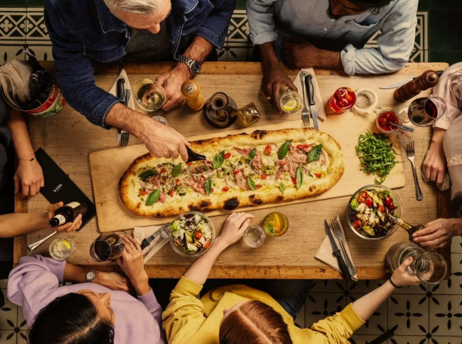 People sitting around a wooden table sharing a meal, including a long pizza and salads, with drinks and condiments.