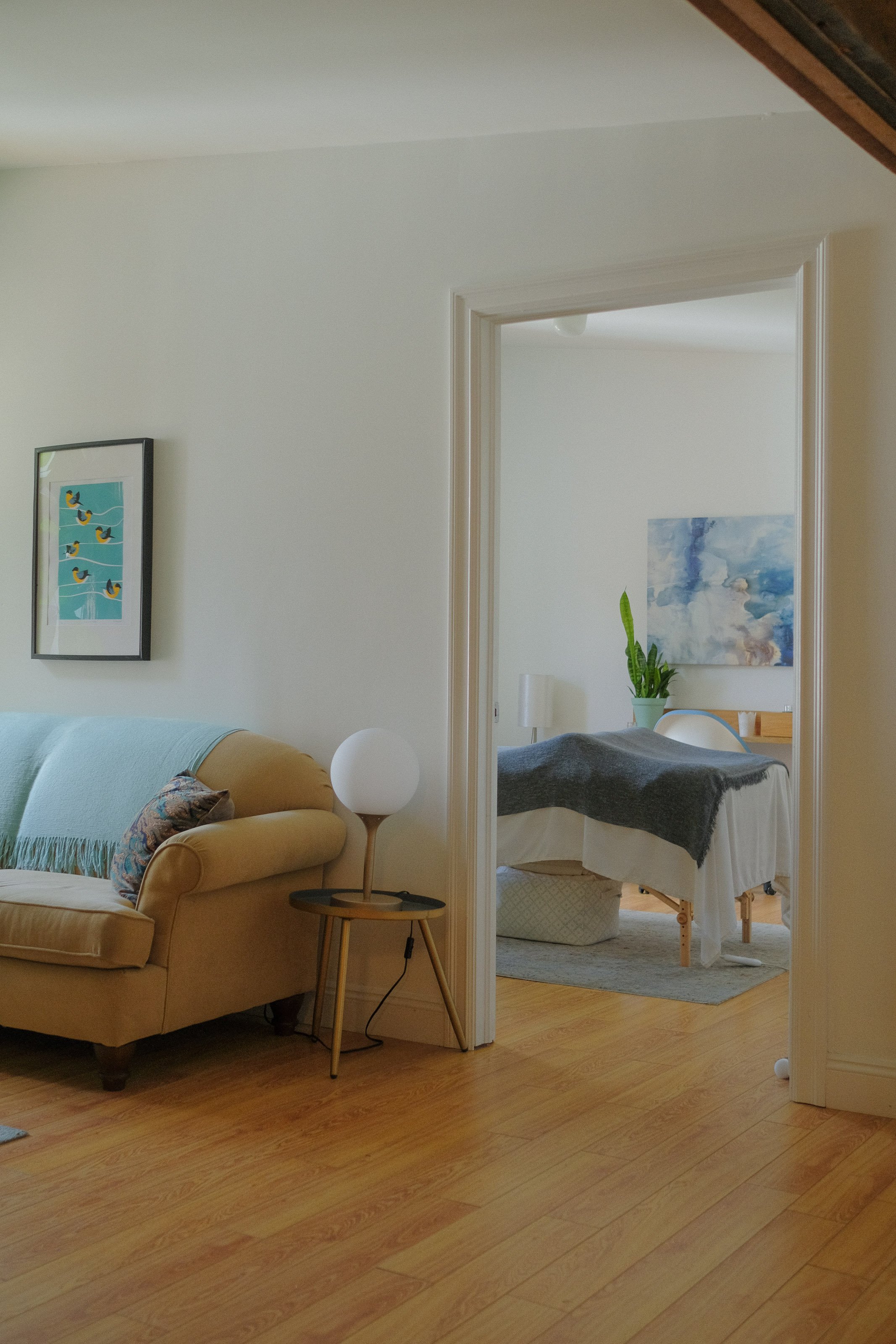Living room with beige armchair, white lampshade, artwork, and hardwood flooring, visible through an open doorway leading to a bedroom with a bed covered with a dark blanket and a large abstract painting on the wall.