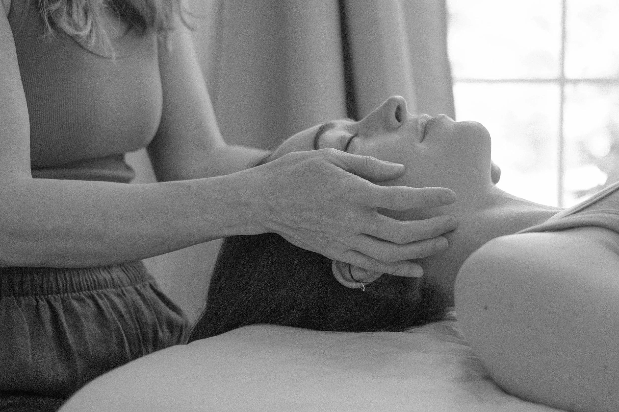 A person lying on their back in a relaxed position with eyes closed while another person holds their head with both hands, possibly receiving a massage.