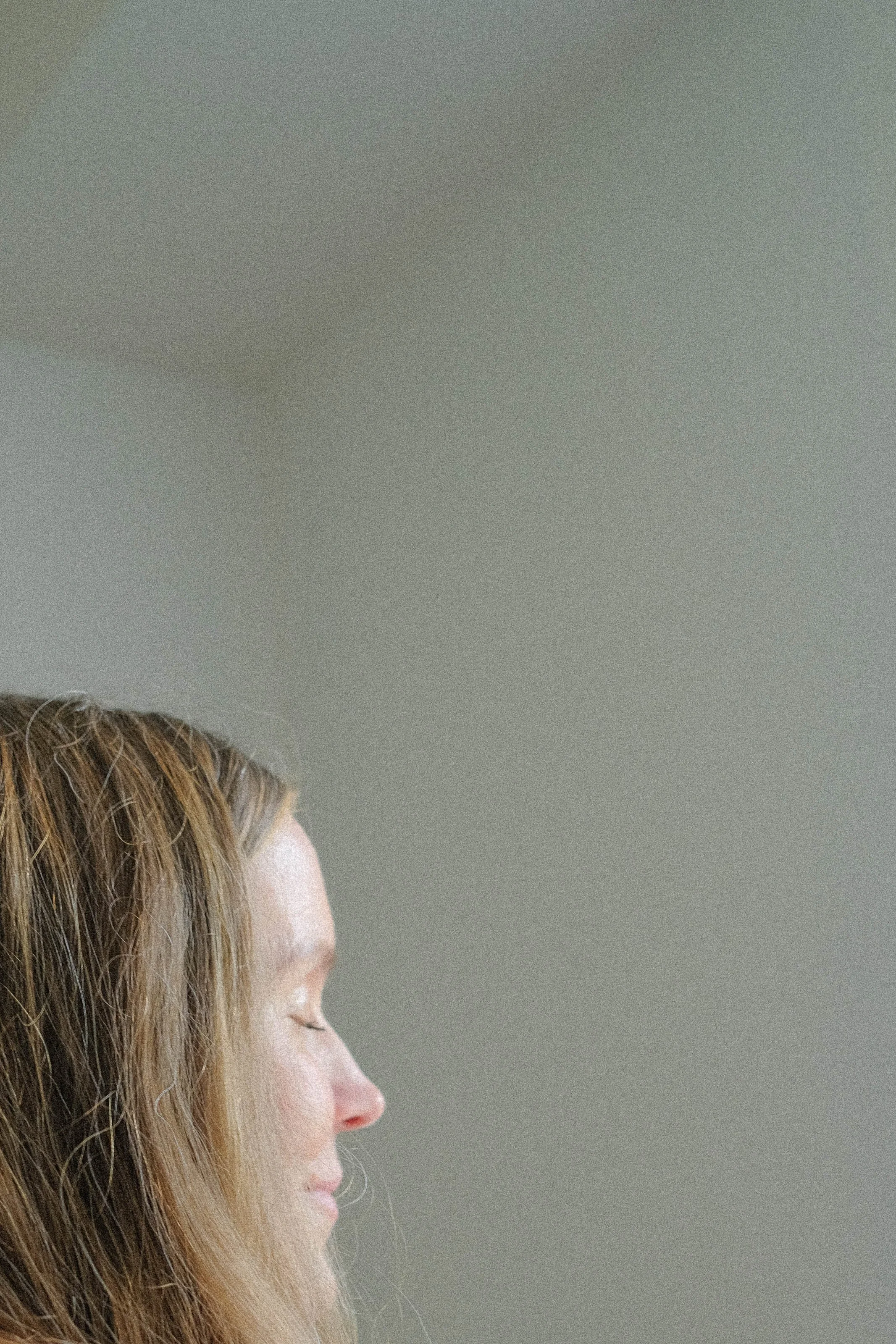 Close-up of a woman's face in profile with closed eyes and light brown hair, against a plain, gray background.