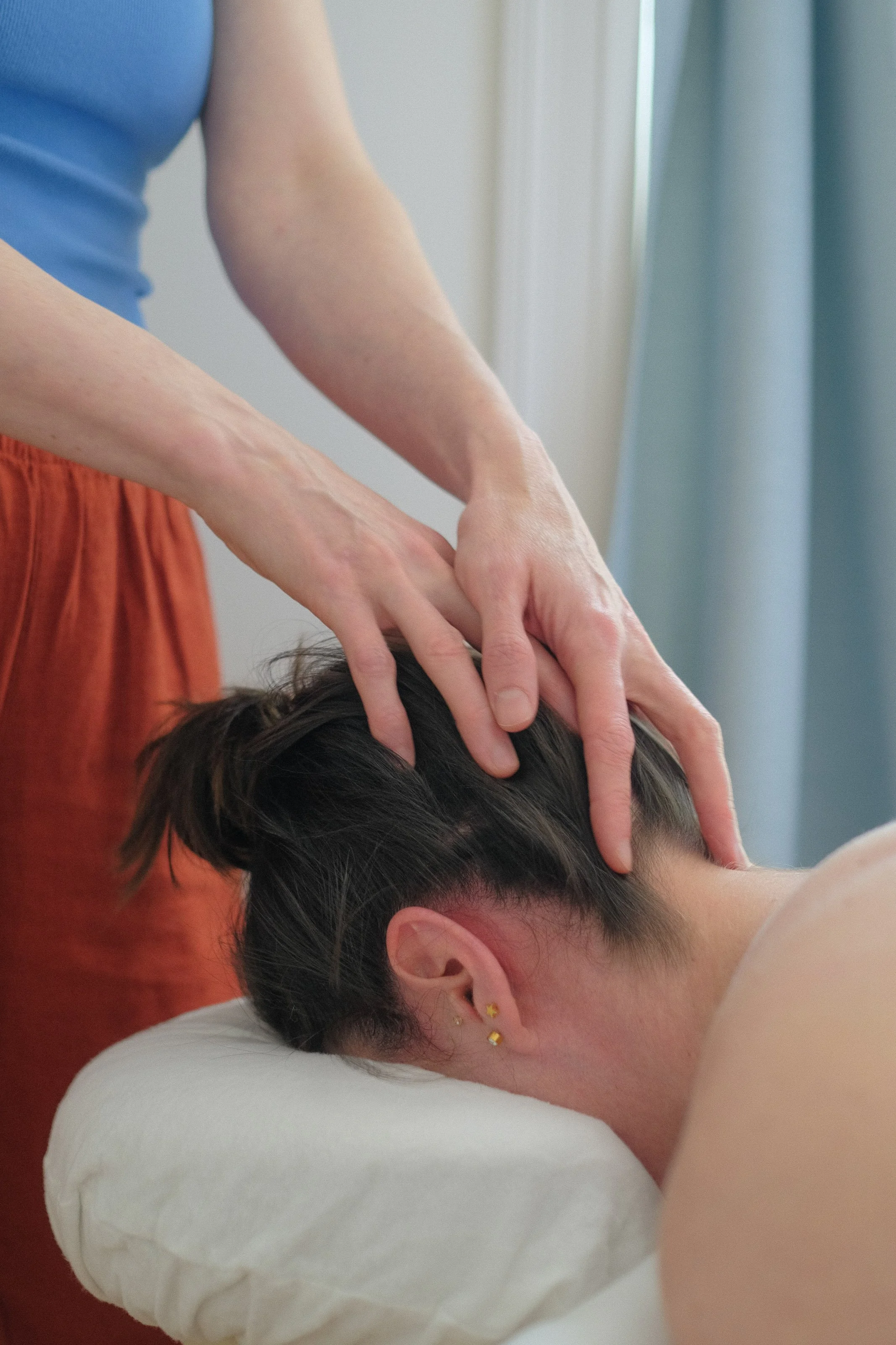 A person receiving a head massage from a massage therapist, who is wearing an orange top. The person on the table has dark hair and is wearing earrings with star-shaped charms. The setting appears to be indoors with a light curtain in the background.
