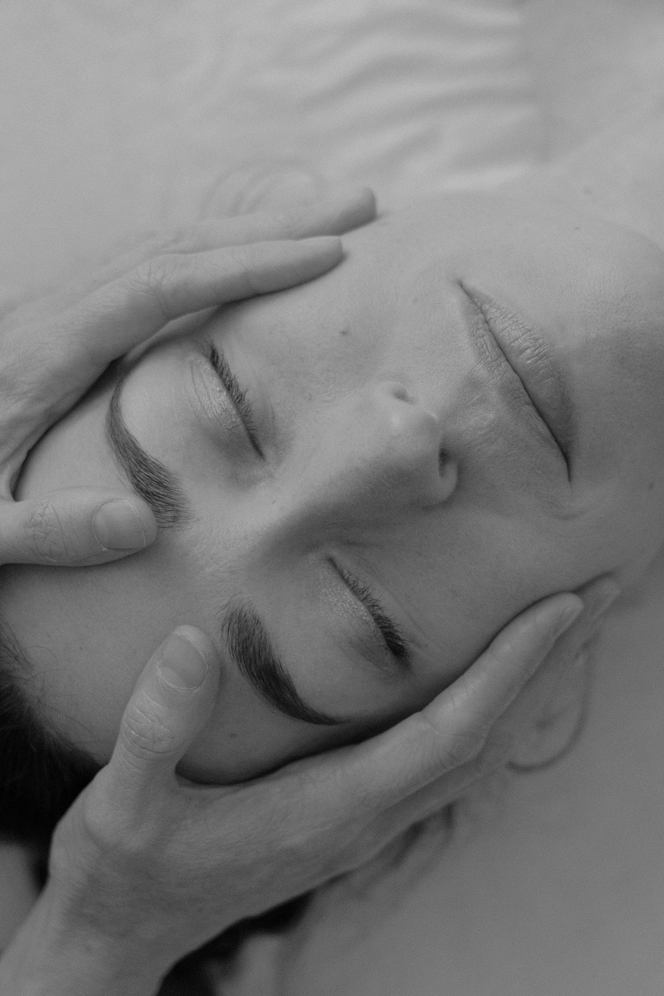 Close-up black and white photograph of a person receiving a facial massage, with hands gently touching their face and eyes closed.