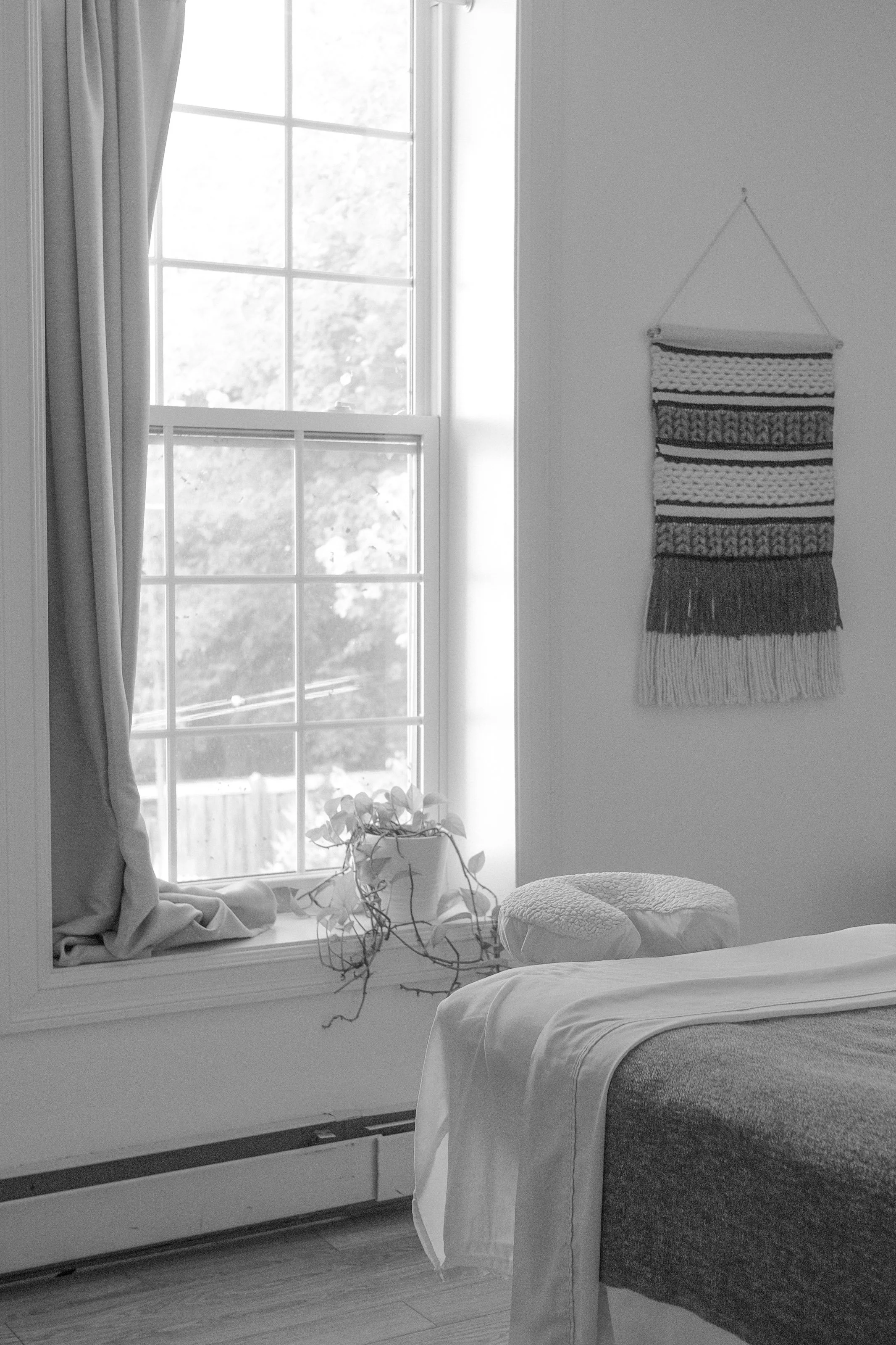 A cozy bedroom features a bed with a white blanket and a small round textured cushion, positioned near a large window with divided panes. The window is partially covered by a runner and a long, light-colored curtain. A potted plant with trailing vines sits on the windowsill. A woven wall hanging with horizontal striped patterns and fringes is displayed on the adjacent wall.