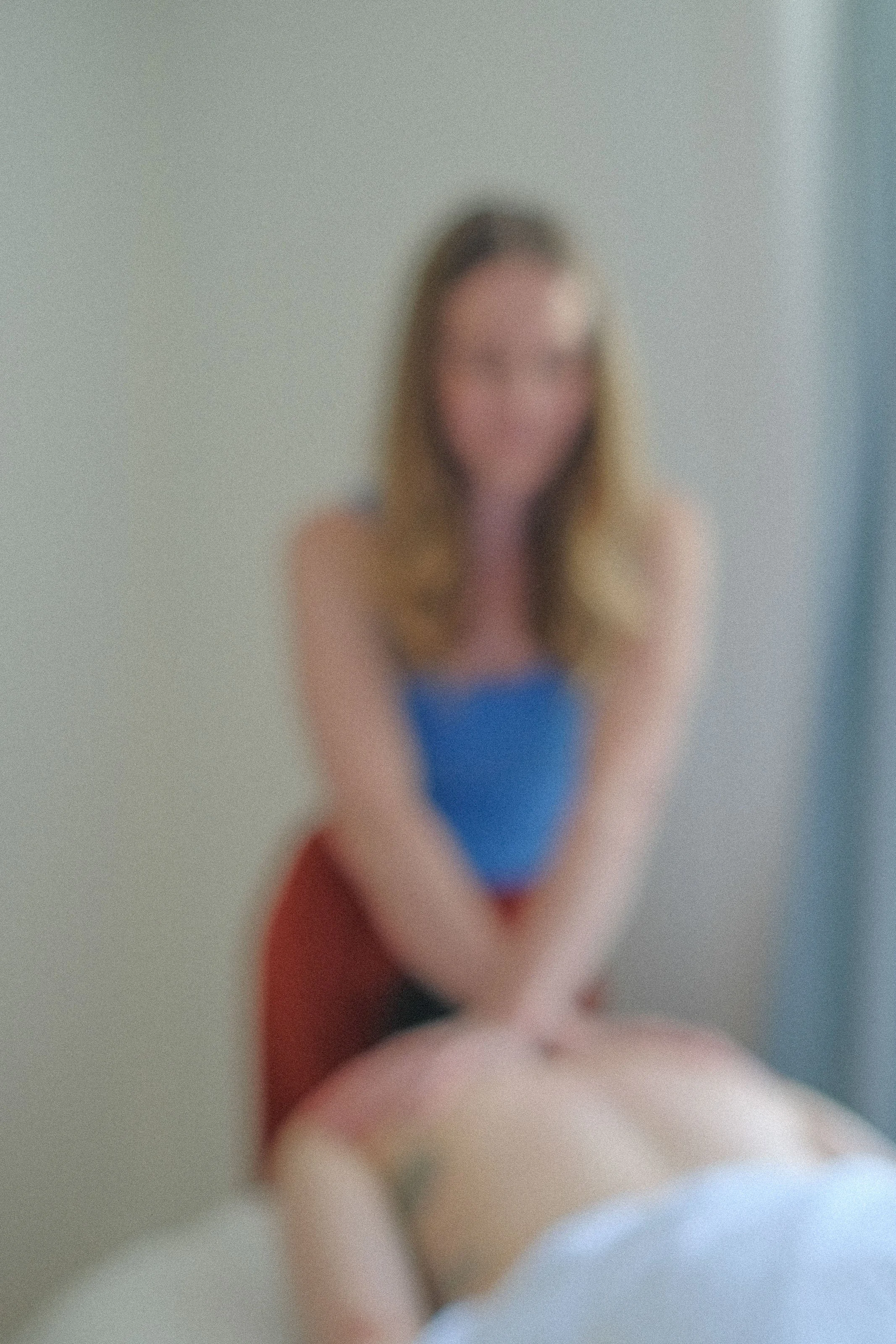 Blurred image of a woman with long hair, wearing a blue top and red bottoms, sitting on the floor against a plain wall.