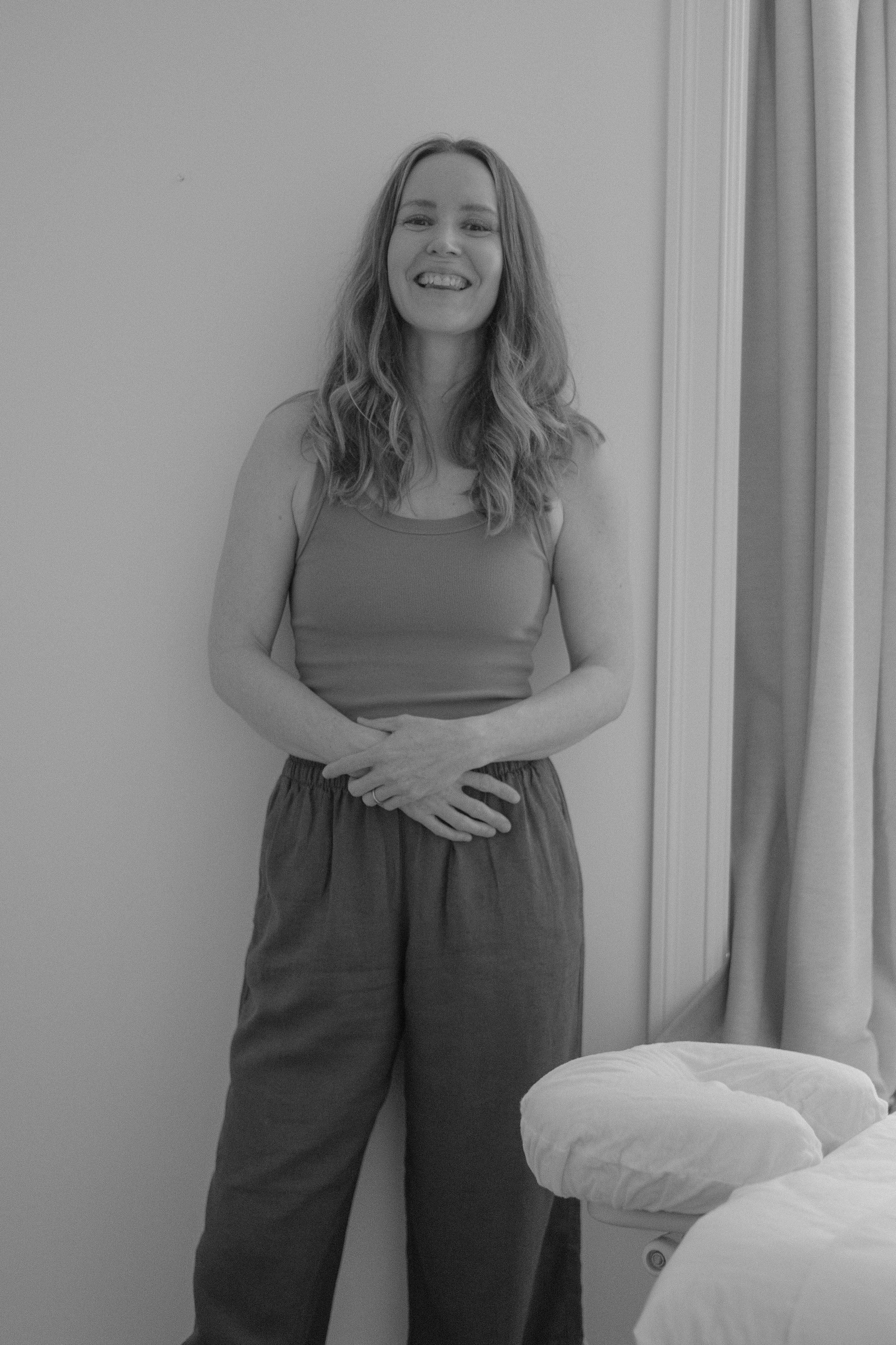 A smiling young woman with long wavy hair standing in a room with plain walls and a bed nearby.