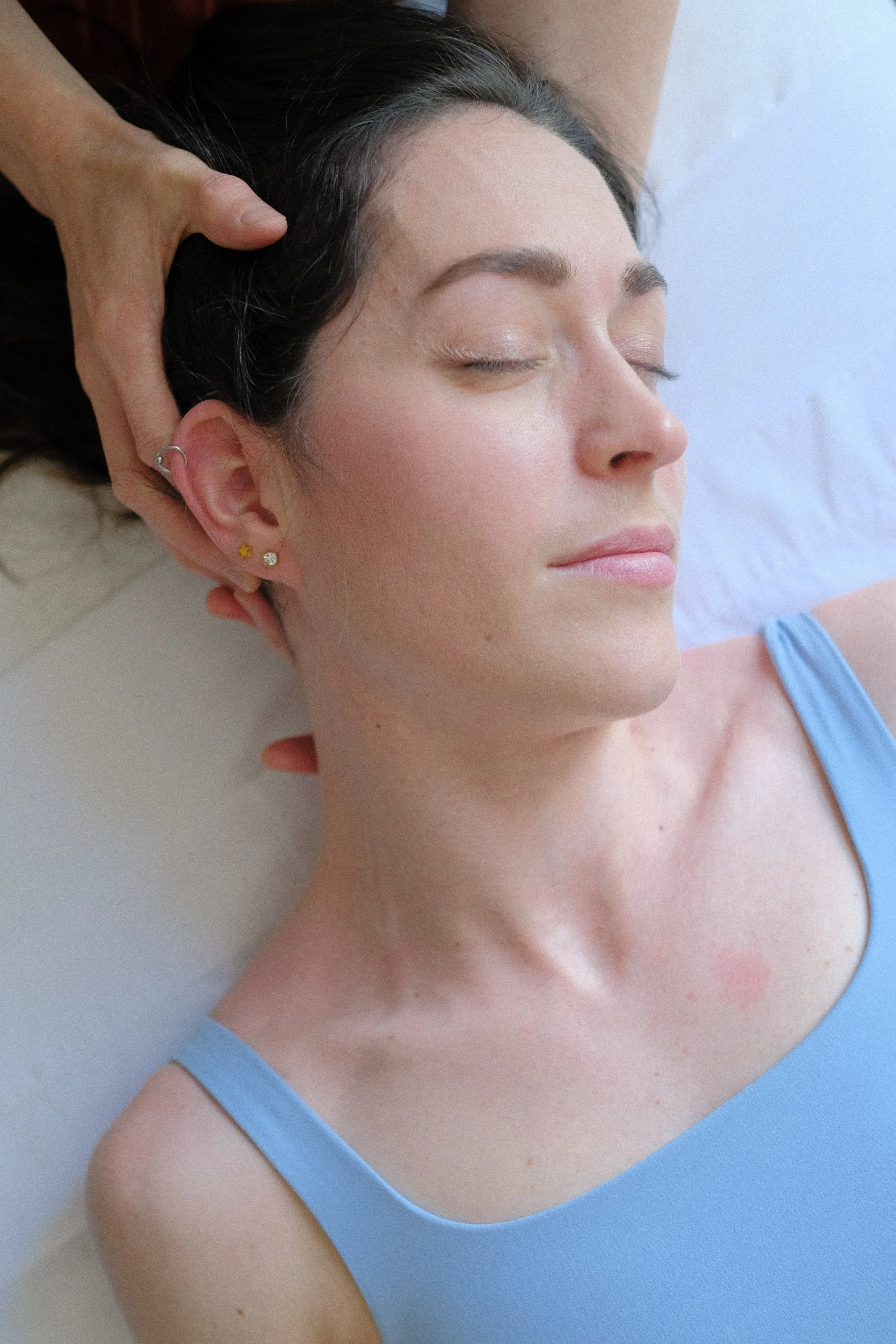 A woman lying on a bed with eyes closed, facial expression peaceful, wearing a blue tank top, getting a massage or facial treatment.