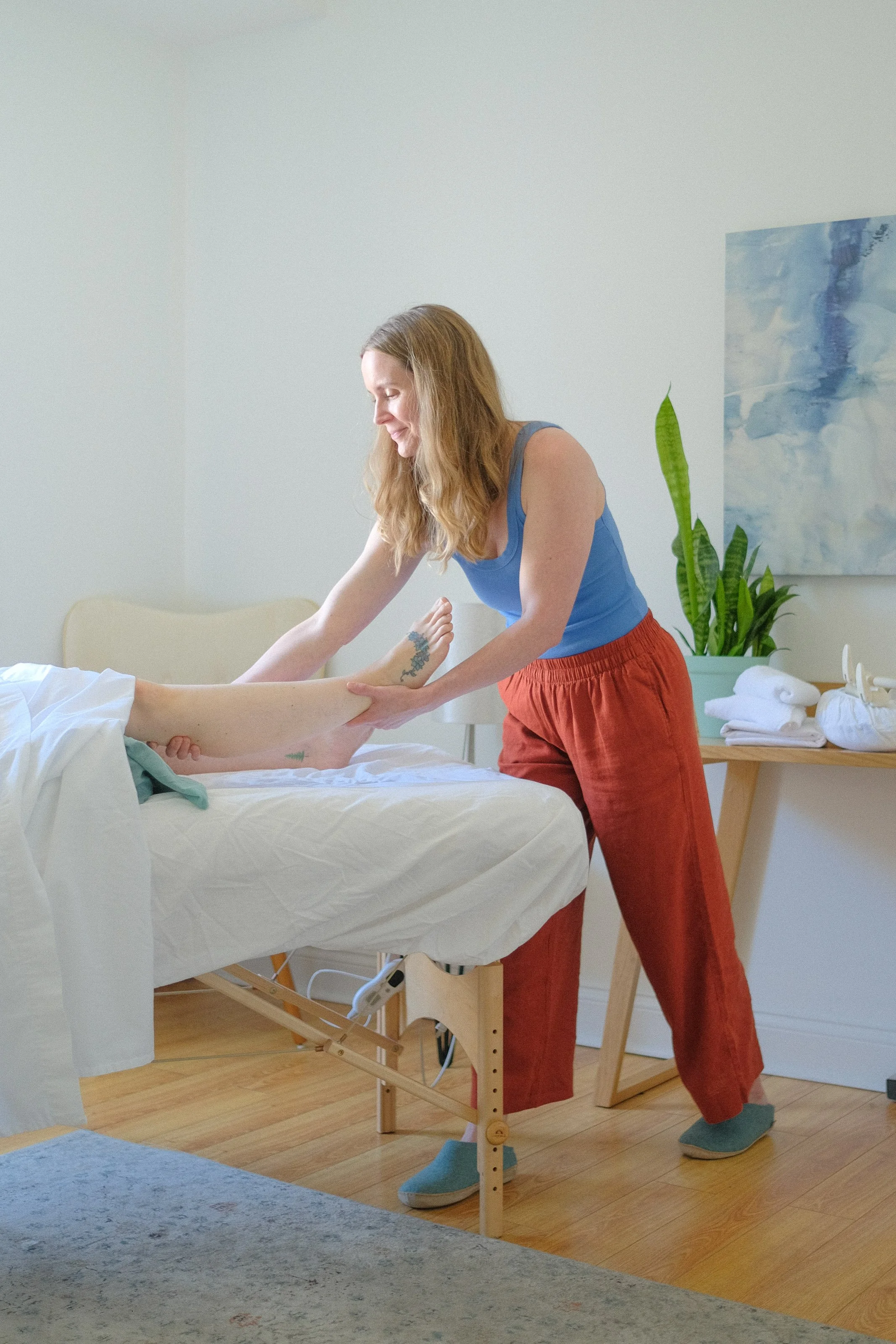 A woman with long, wavy brown hair, wearing a blue tank top and red-orange pants, is standing by a bed and gently holding the leg of a person lying in bed. The setting appears to be a bright, minimalist bedroom with a light-colored wall, a piece of abstract blue and white artwork, a green potted plant, and a wooden table with neatly folded white towels.