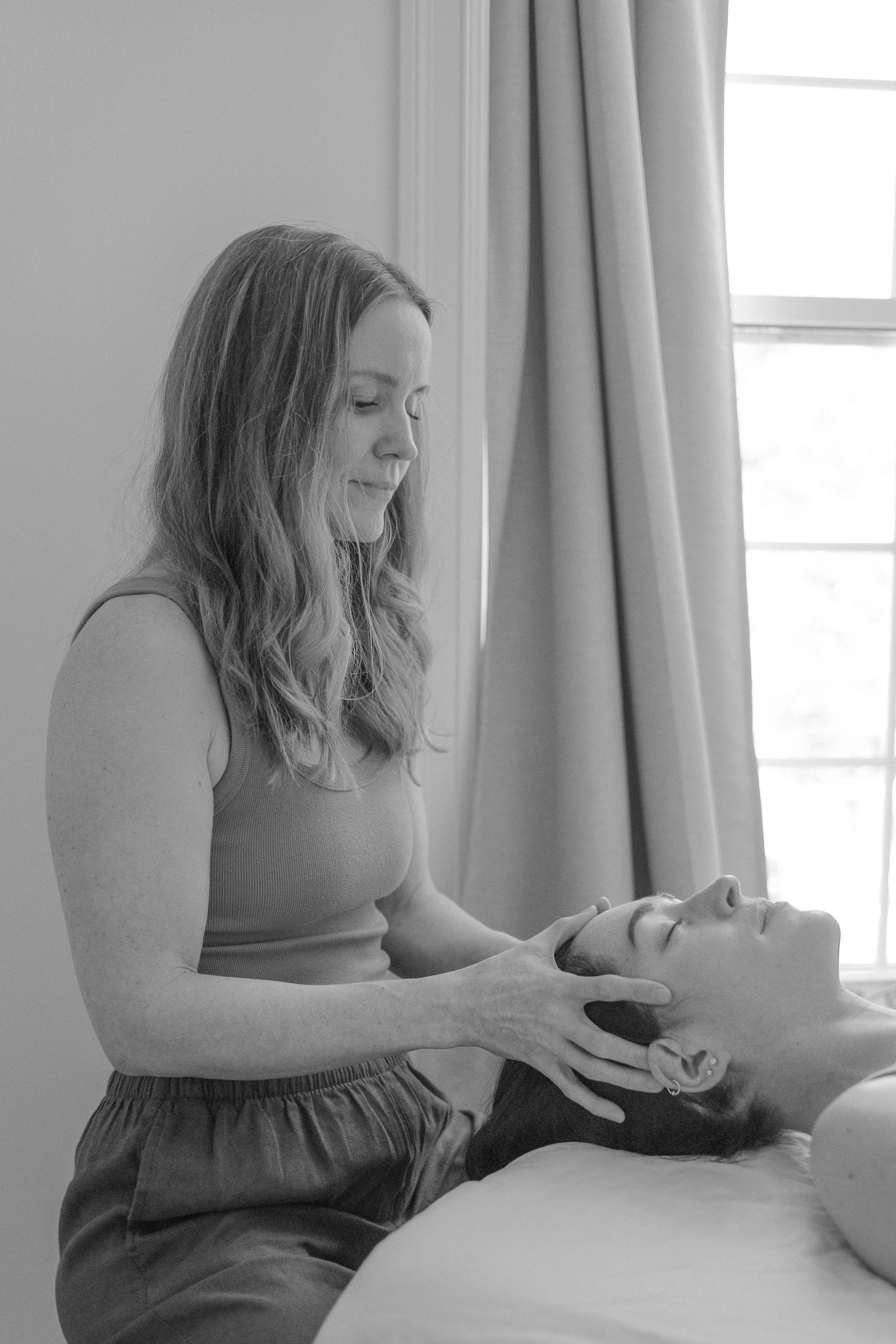 A woman receiving a massage from a massage therapist who appears to be lying on a massage table near a window with curtains.
