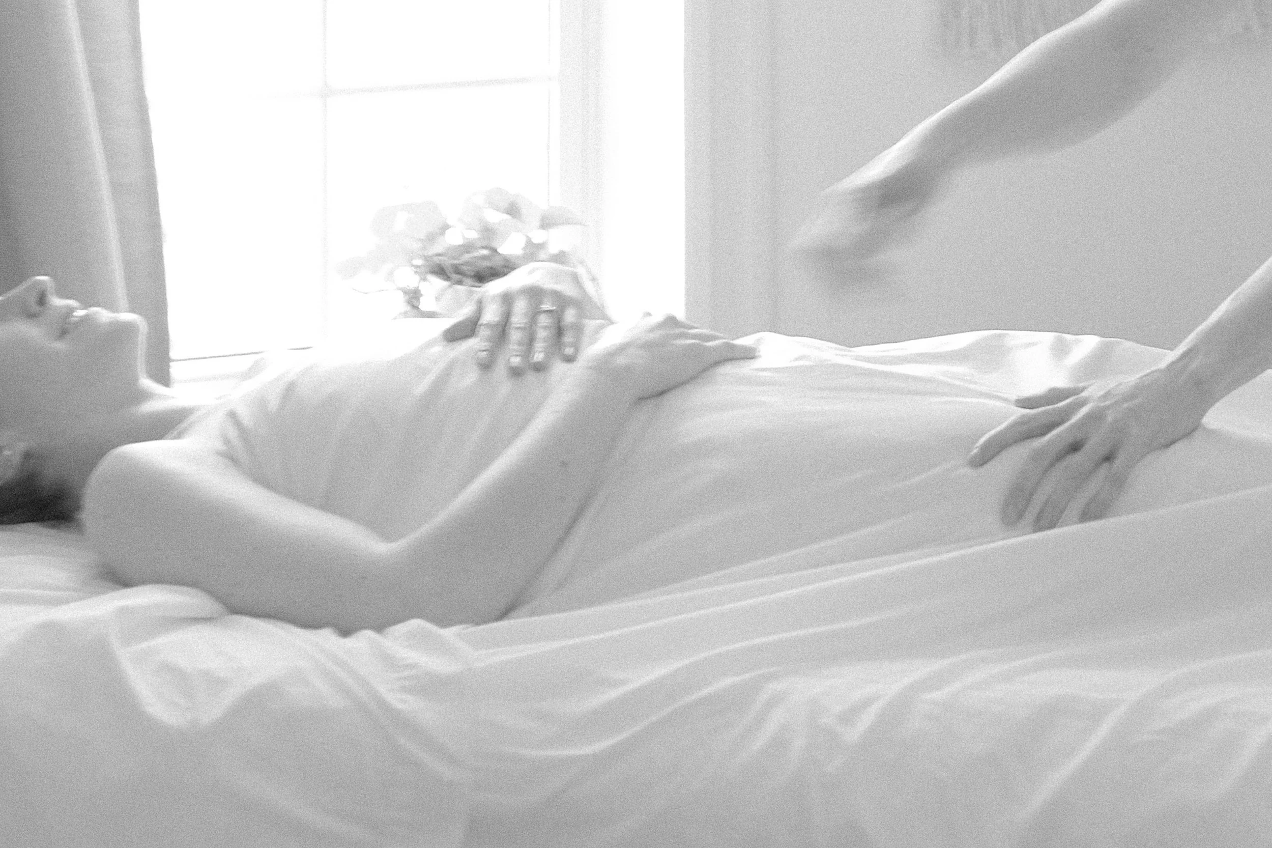 A person lying in bed next to a window, with the sunlight illuminating the room, capturing part of their face and hand resting on their chest.