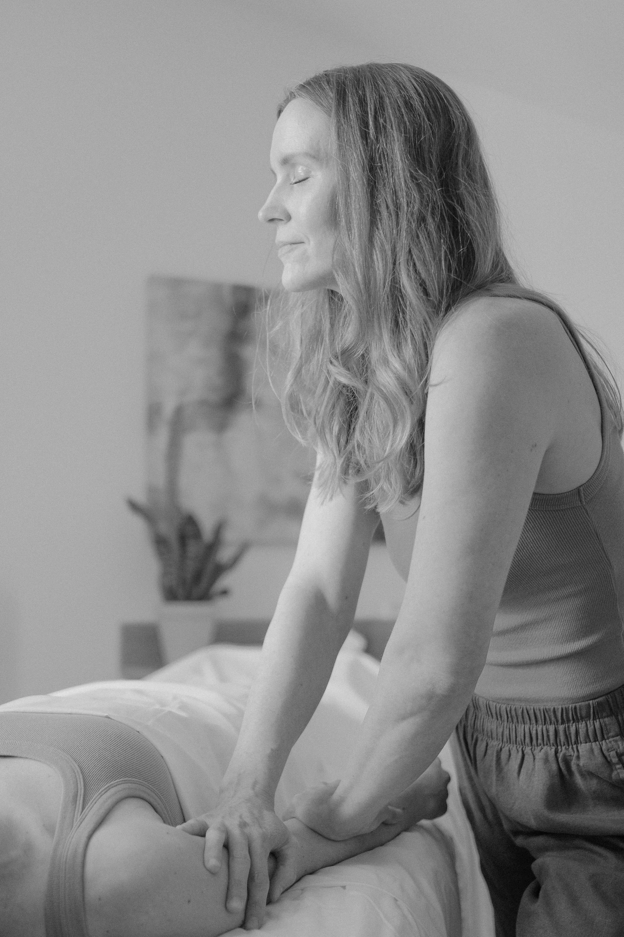 A woman performing a massage on a person lying face down, with her eyes closed and a peaceful expression. The woman has long wavy hair and is wearing a sleeveless top. The setting appears to be a spa or wellness center.