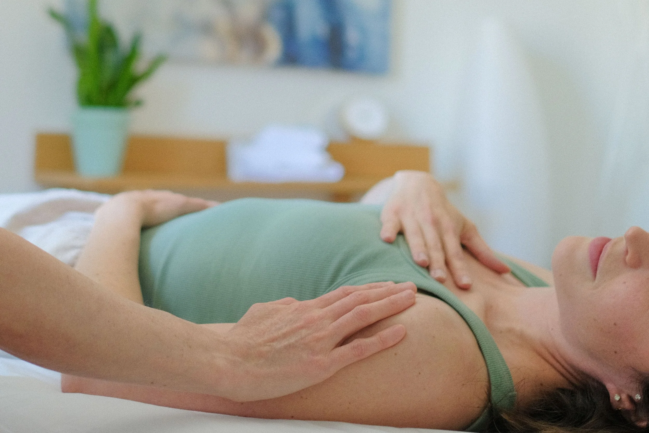 Person receiving a massage or physical therapy, lying on a massage table with hands on their back, in a bright room with a plant and a painting on the wall.