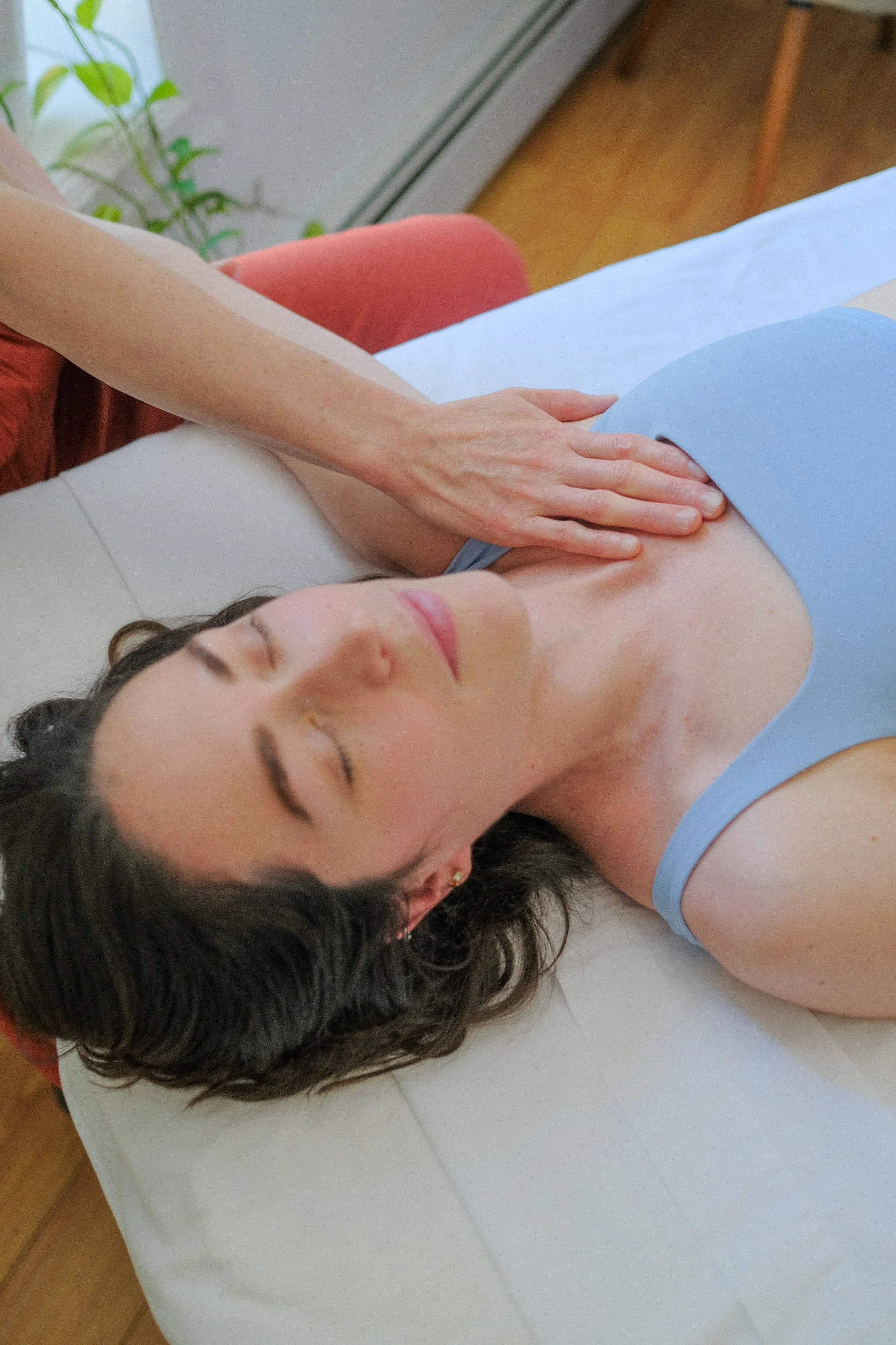 A woman lying on her side on a massage table with her eyes closed, receiving a massage, in a room with a window and a plant.