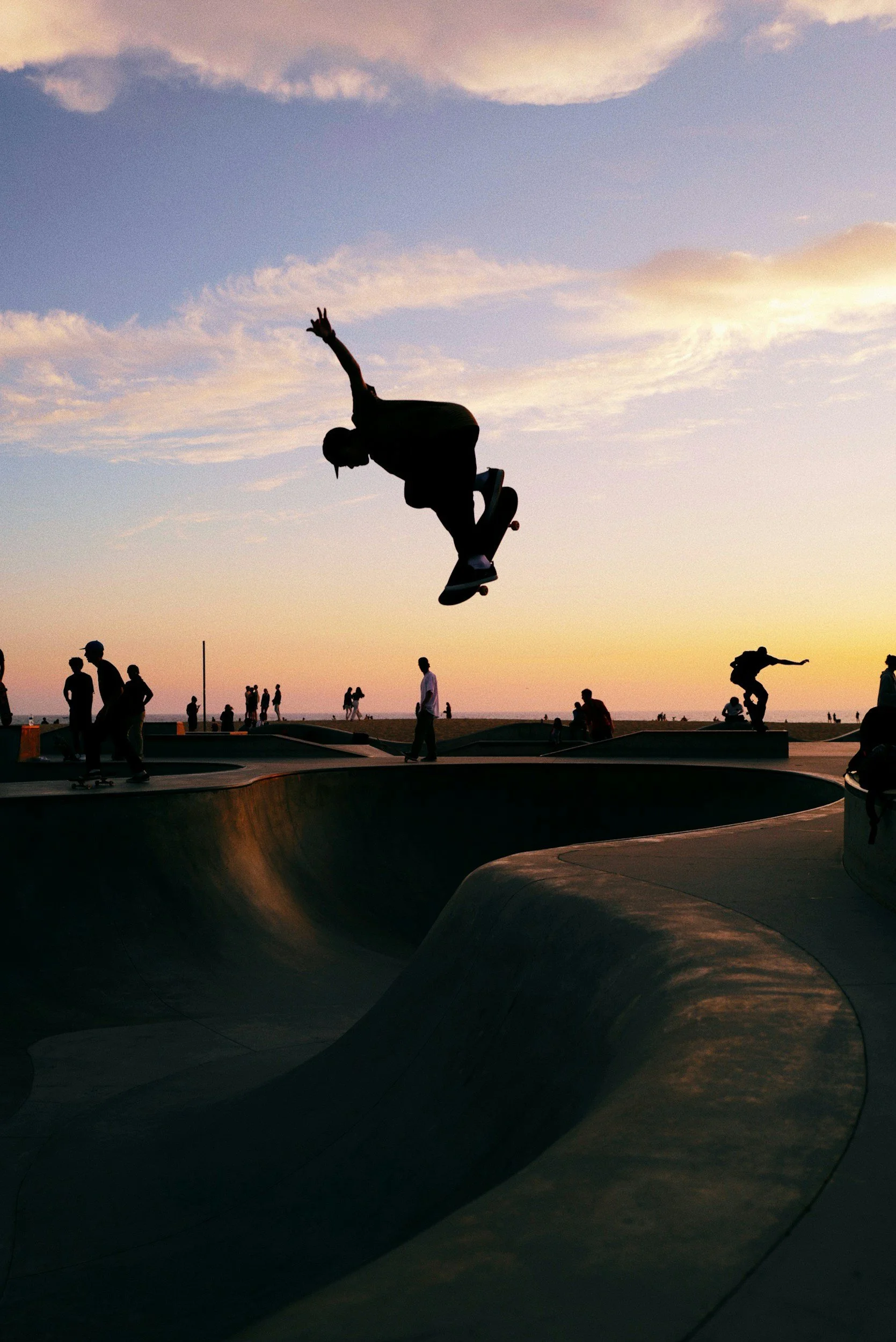 Silhouettes of skateboarders performing tricks at a skatepark during sunset, with a cloudy sky and people in the background.