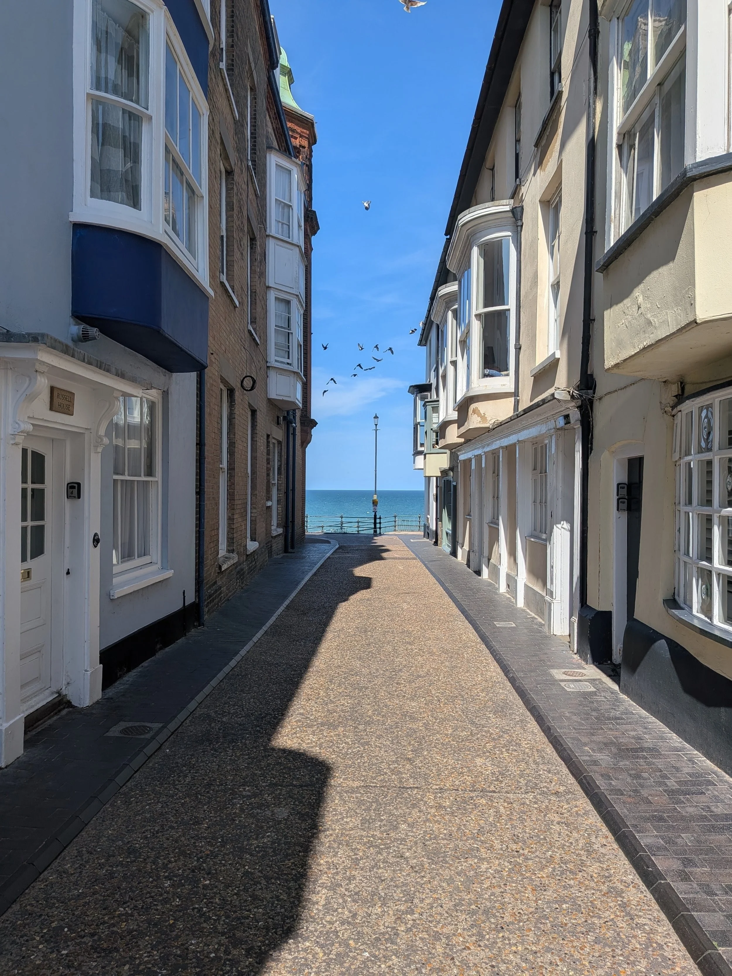 A narrow alleyway between two buildings opening towards the seaside, with a streetlamp in the distance, birds flying in the blue sky.