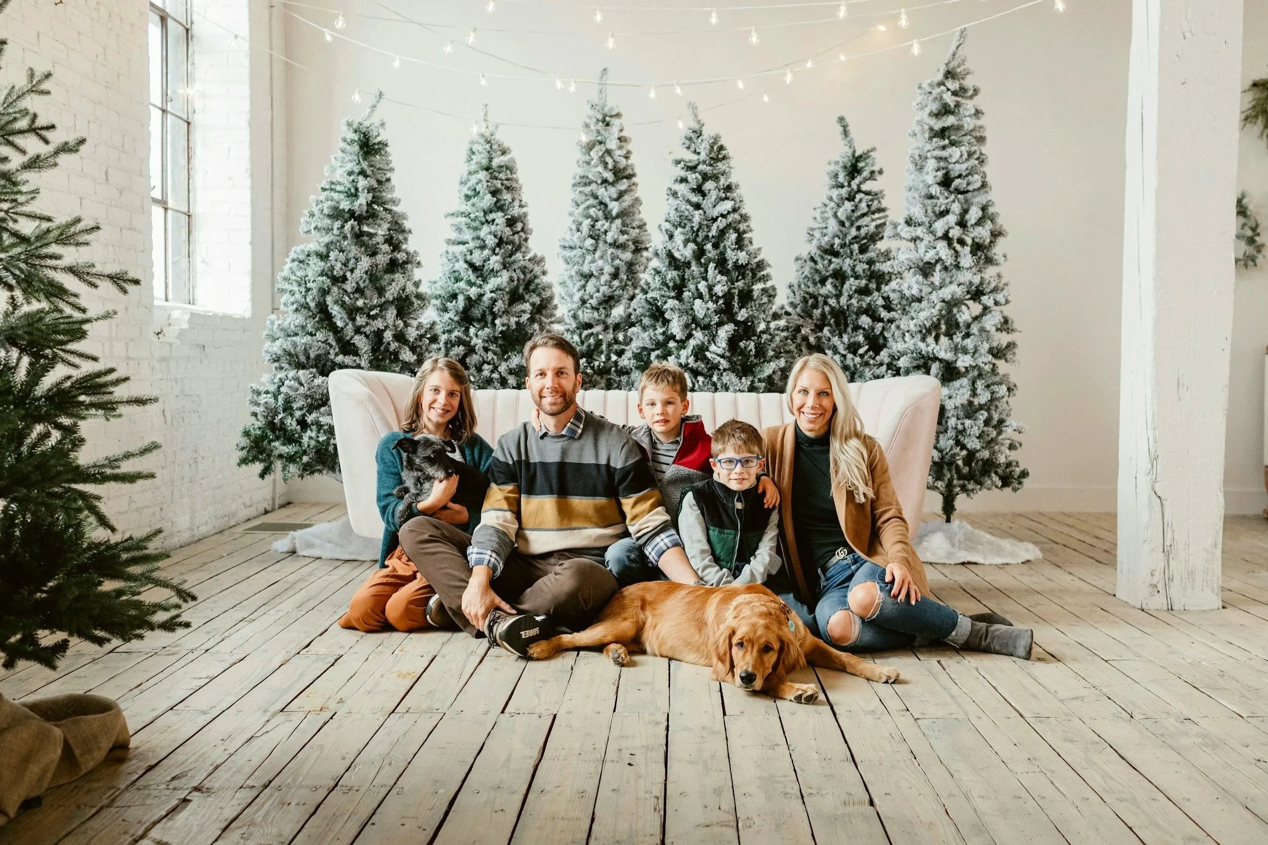 A family of five with a dog sitting on a wooden floor in front of a white tufted sofa with snow-covered Christmas trees behind them. The family includes a woman, a man, two young boys, and a girl, all smiling.