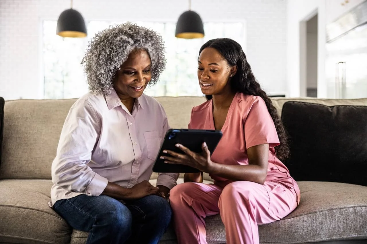 Two women sitting on a beige sofa in a well-lit room, looking at a tablet together. One woman has gray curly hair and is wearing a light pink shirt, and the other woman has long dark hair and is wearing pink scrubs.