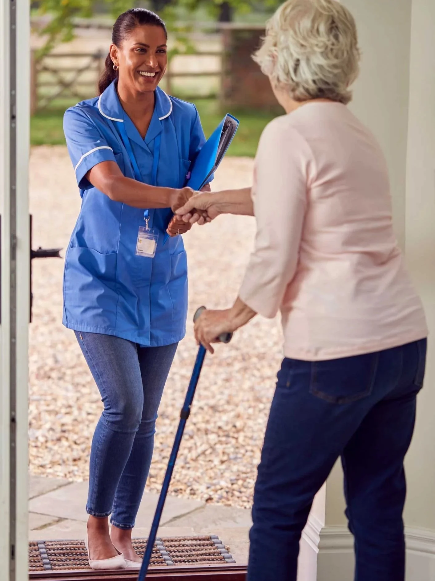A nurse in blue scrubs smiling and shaking hands with an elderly woman at the doorway of her house, with the woman using a cane.