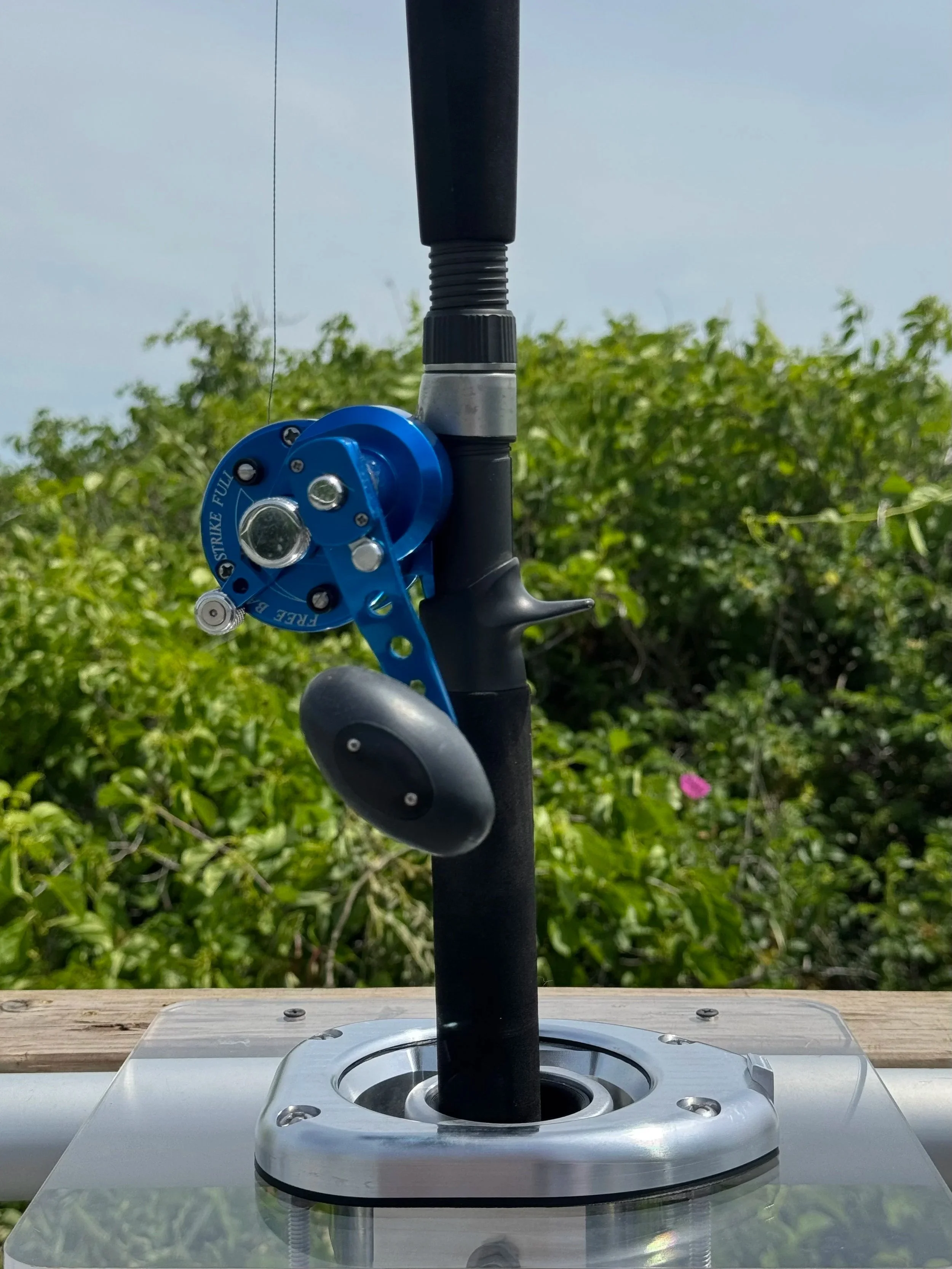 This is a close-up photo of a black and blue robotic device with a circular component labeled 'Strike Full.' It is mounted on a metallic base, and a black pole extends upward from the base. The background features greenery and a bright blue sky.