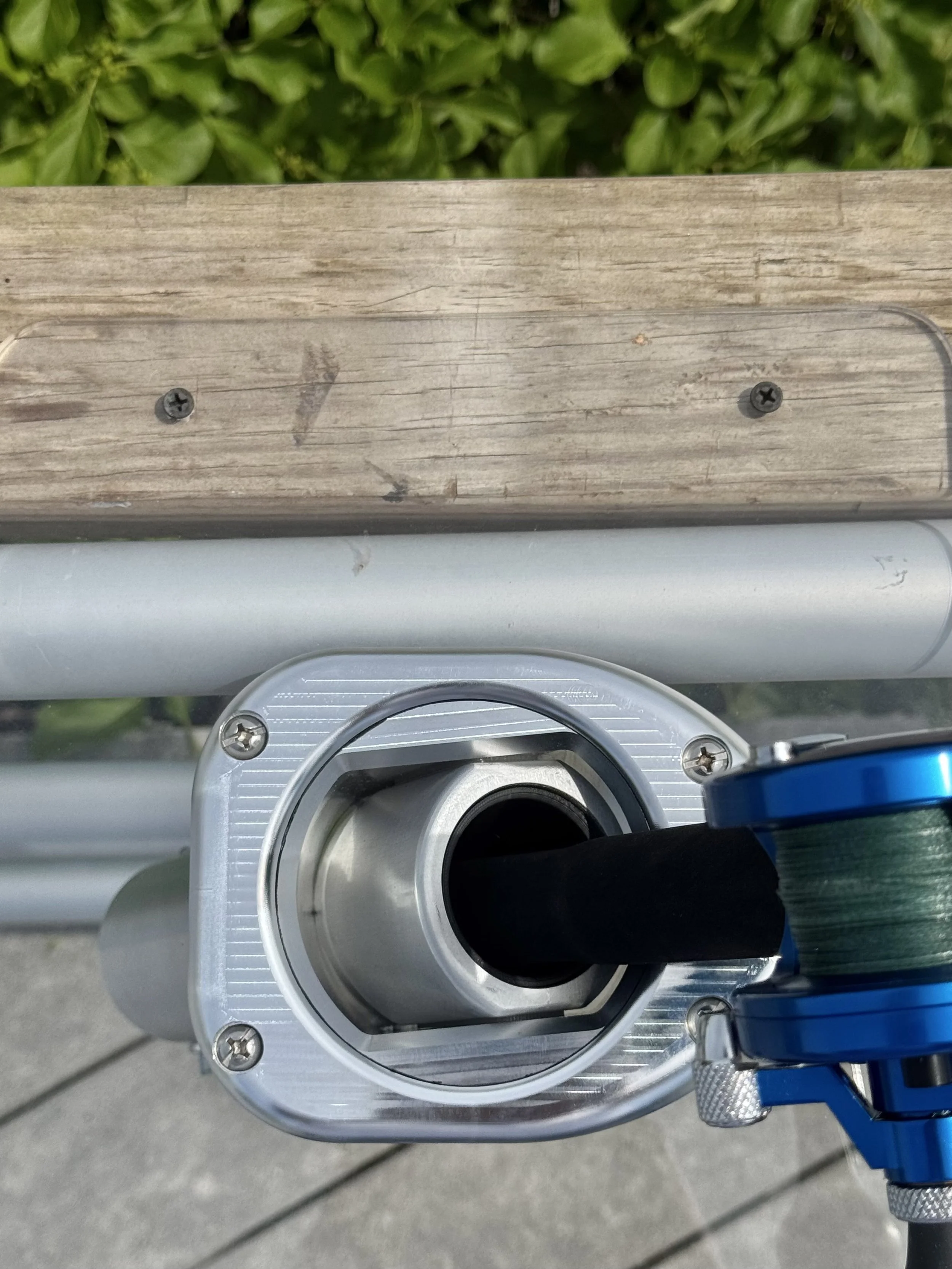 Close-up of a metal and plastic valve with a black handle, mounted on a wooden surface with a screw, with green foliage in the background.