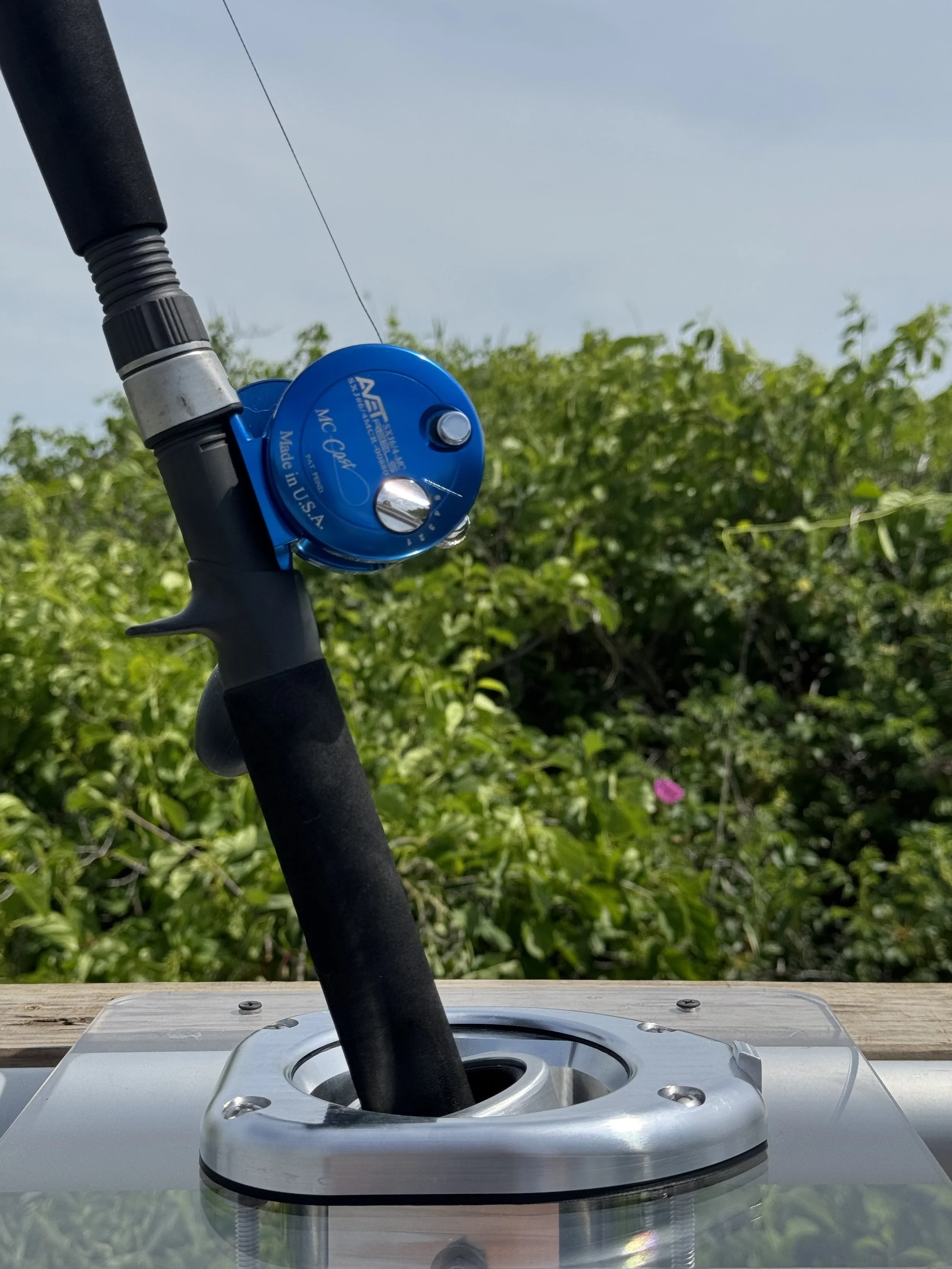 Close-up of a blue AFNMC fishing reel mounted on a black fishing rod, sitting in a metallic stand outdoors with green bushes and a cloudy sky in the background.