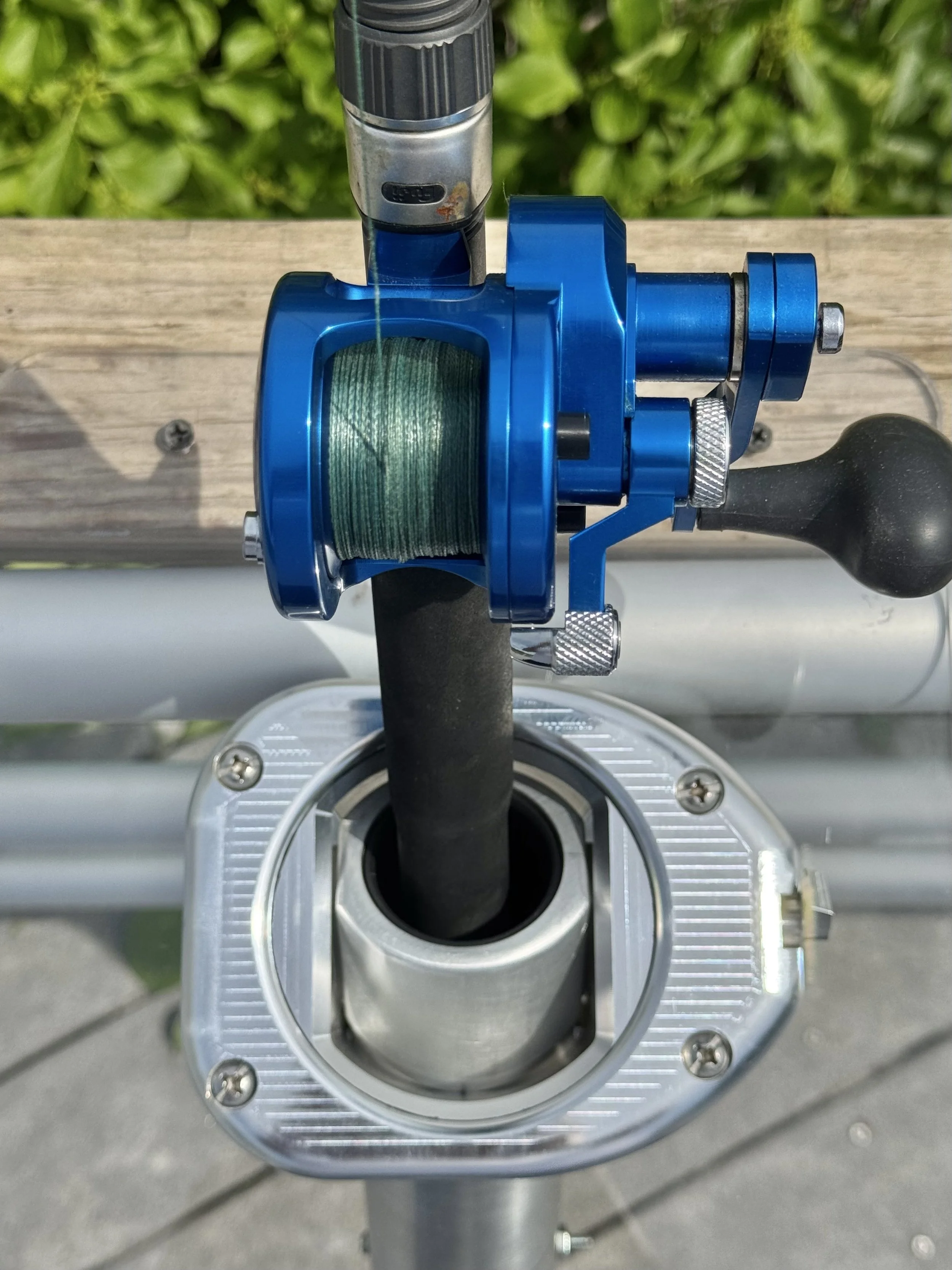 Close-up of a purple fishing reel attached to a fishing rod, mounted on a metallic stand, with greenery in the background.