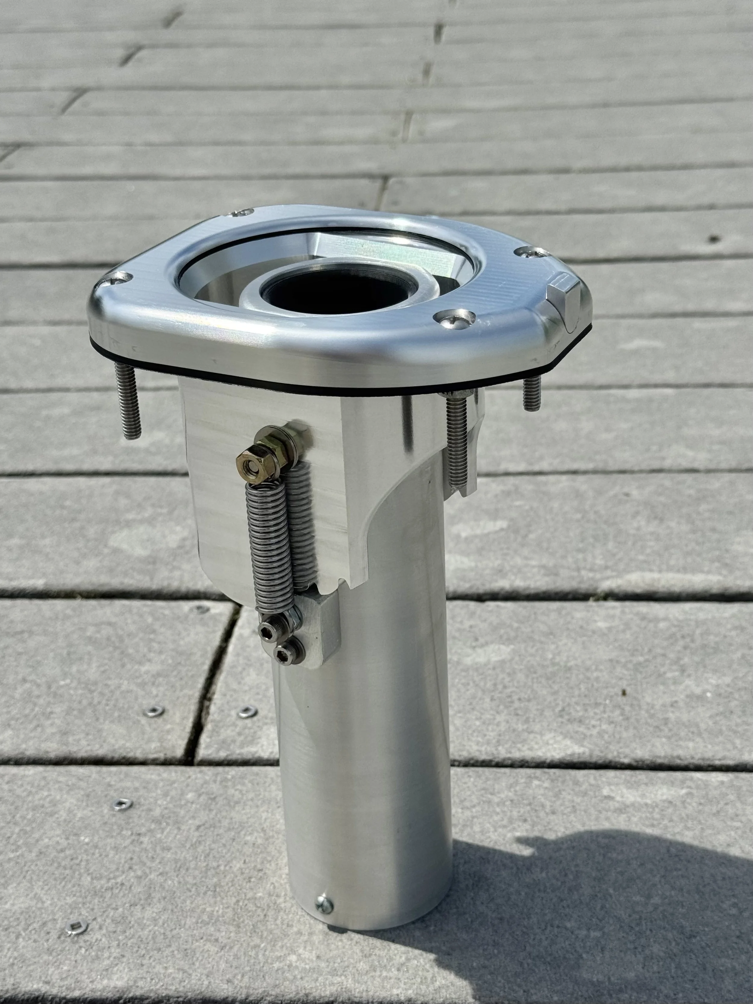 Close-up of a metallic mechanical component with a circular opening, mounted on a cylindrical stand with springs and bolts, placed on an outdoor wooden deck.