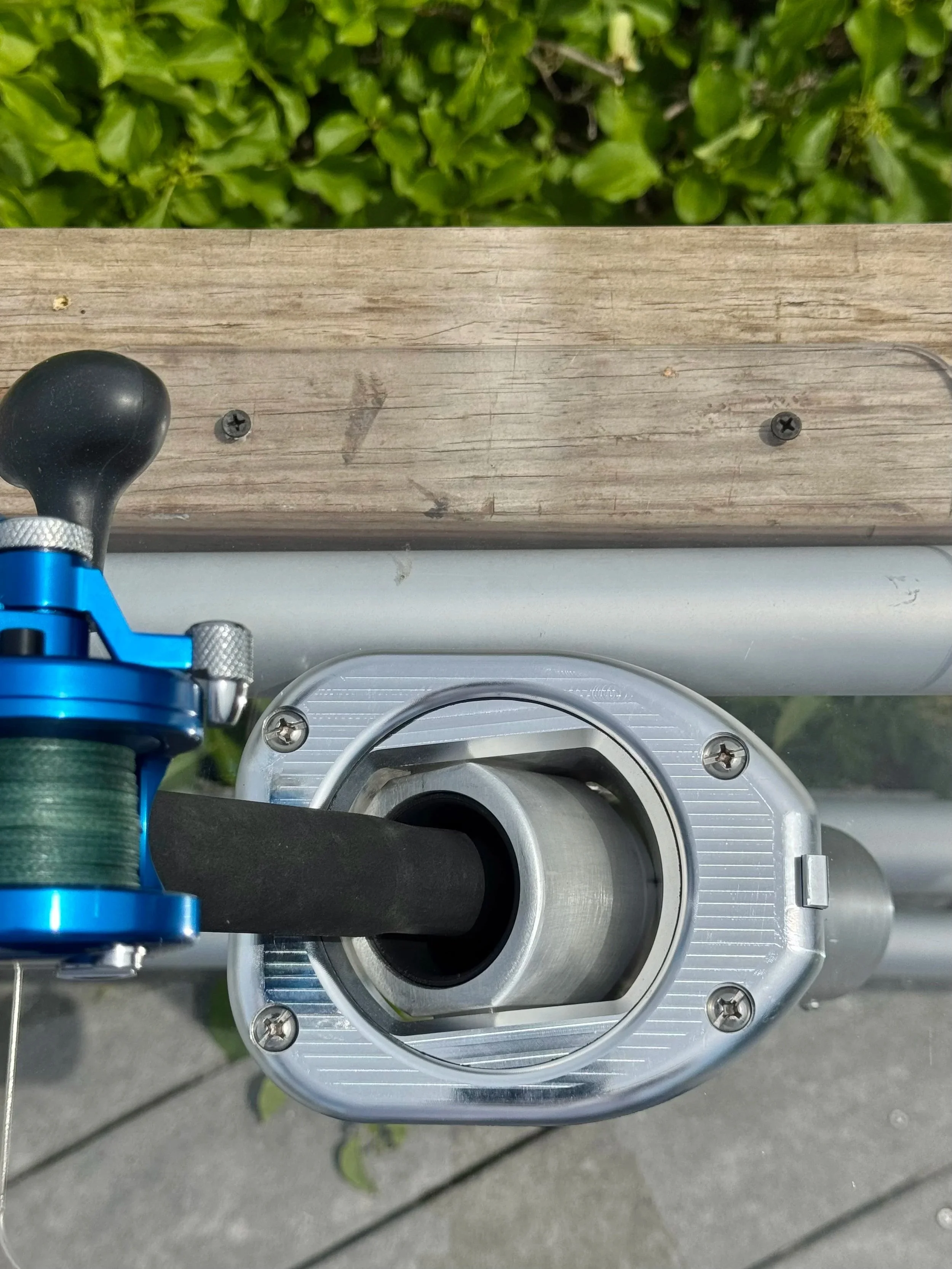 View of a metallic mechanism with a black circular opening, attached to a blue device with screws, against a background of green foliage and a wooden surface.