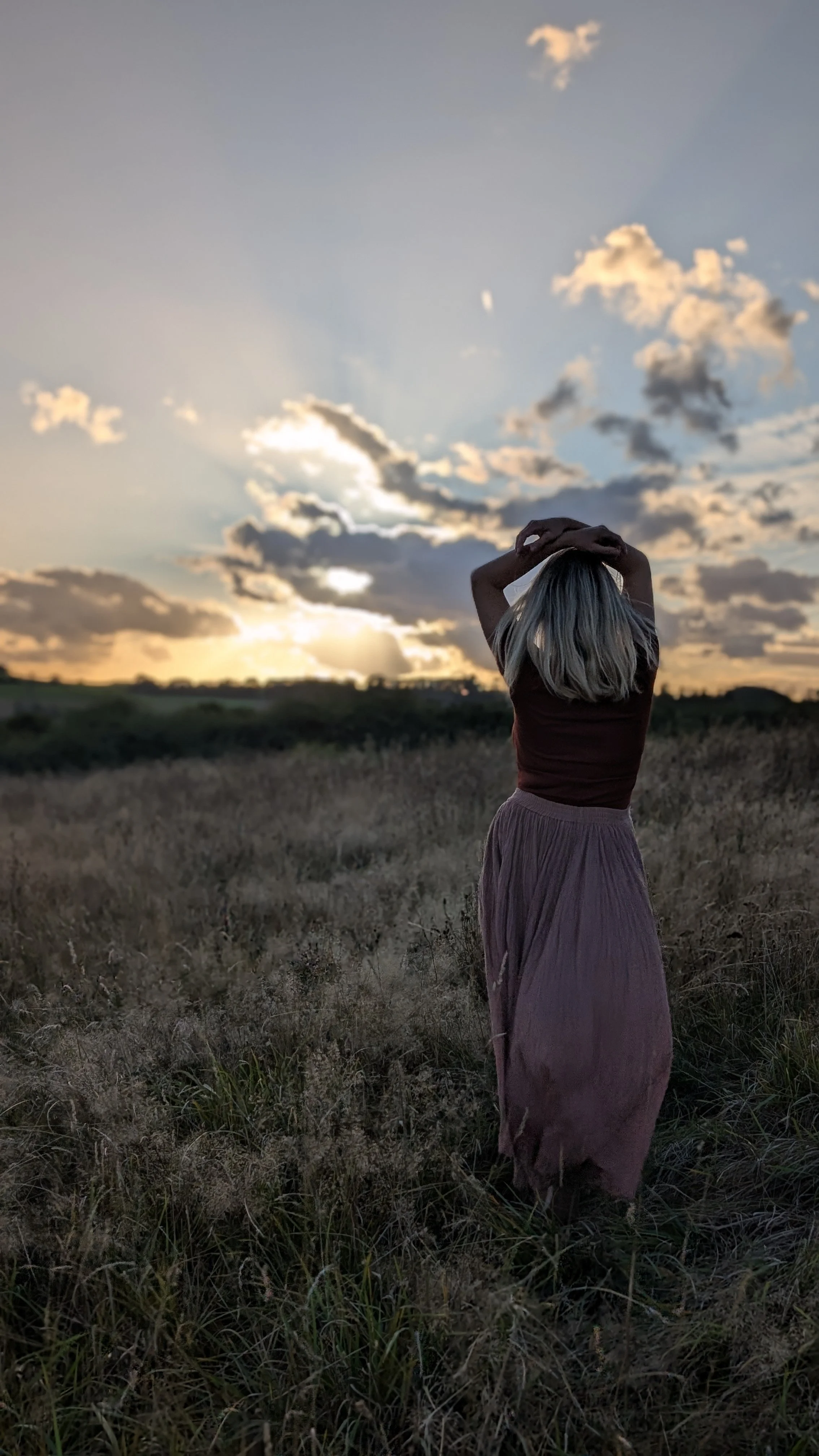 Eine Frau steht in einem Feld bei Sonnenuntergang, blickt in den Himmel und hebt die Arme über den Kopf, während Wolken und das Licht untergehen.