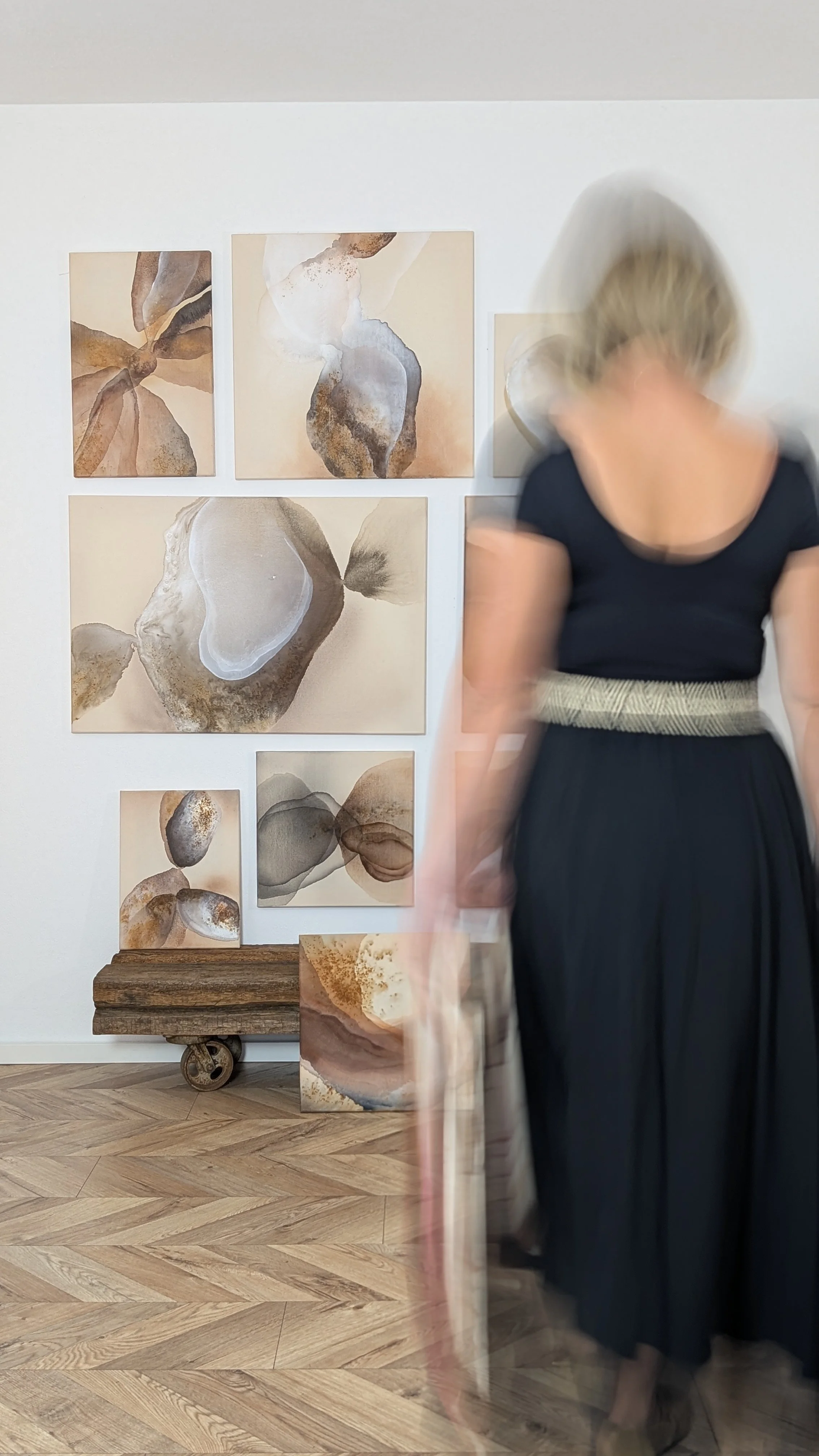Eine Wand mit abstrakten Kunstwerken in Erdtönen, daneben eine Frau in schwarzem Kleid und Tuch, die langsam vorbeigeht.