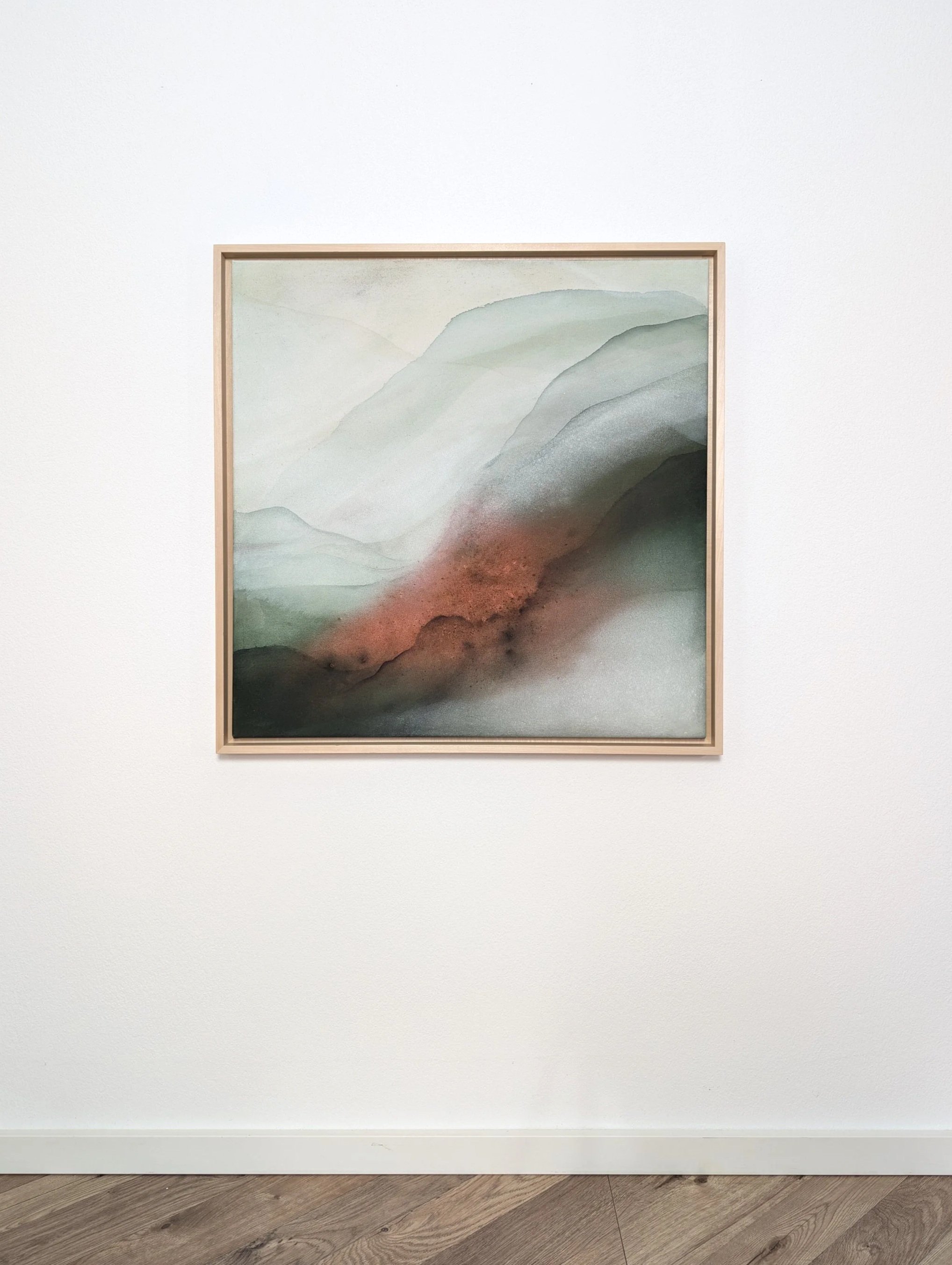 Abstraktes Aquarell-Gemälde mit sanften Grüntönen, Grautönen und einem Hauch von Braun in einem schlichten Holzrahmen. Das Bild ist an einer weißen Wand in einem Museum oder einer Galerie aufgehängt.