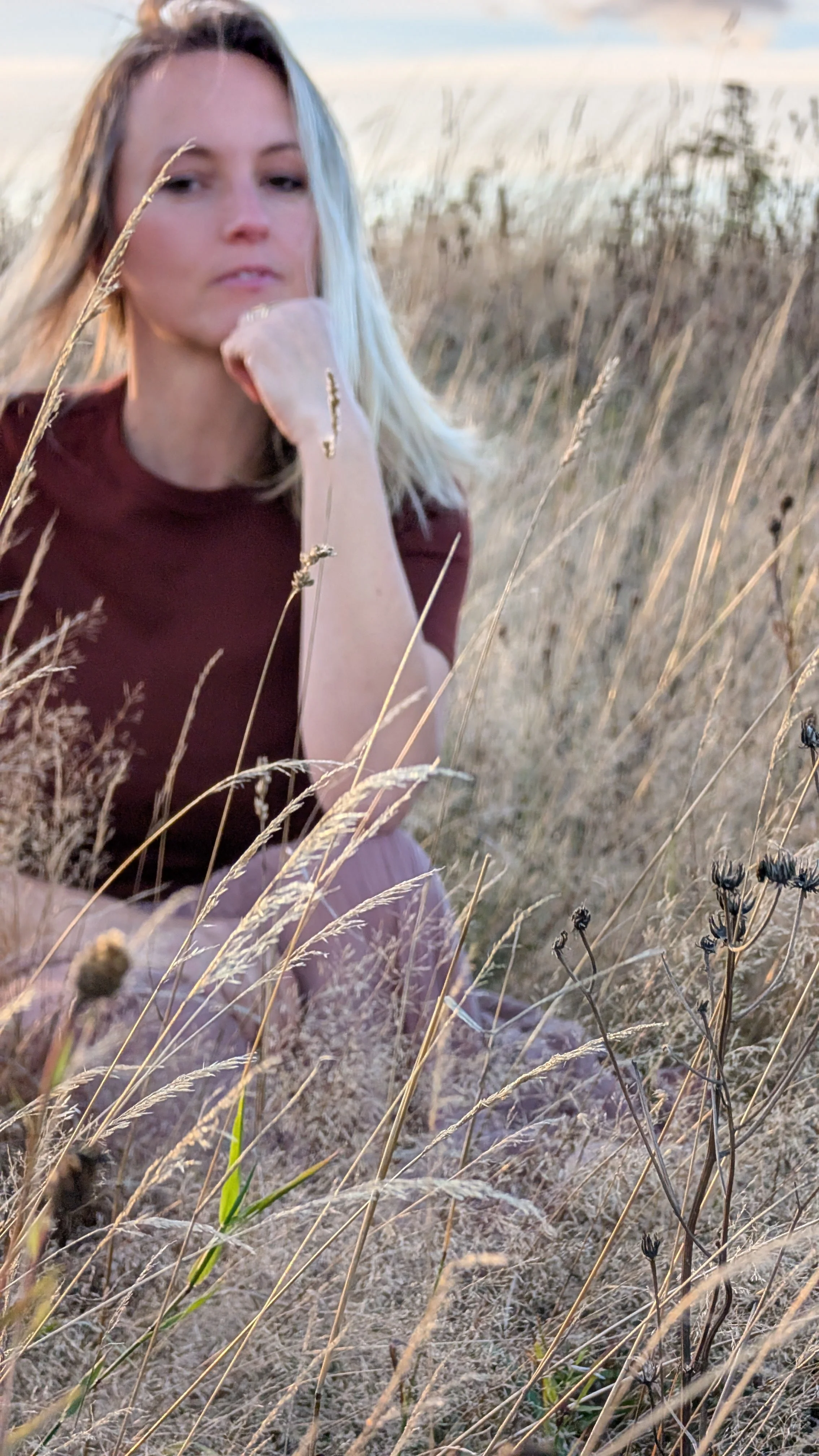 Eine junge Frau mit blonden Haaren, die in einem Feld mit trockenen Gräsern sitzt und nachdenklich aussieht.
