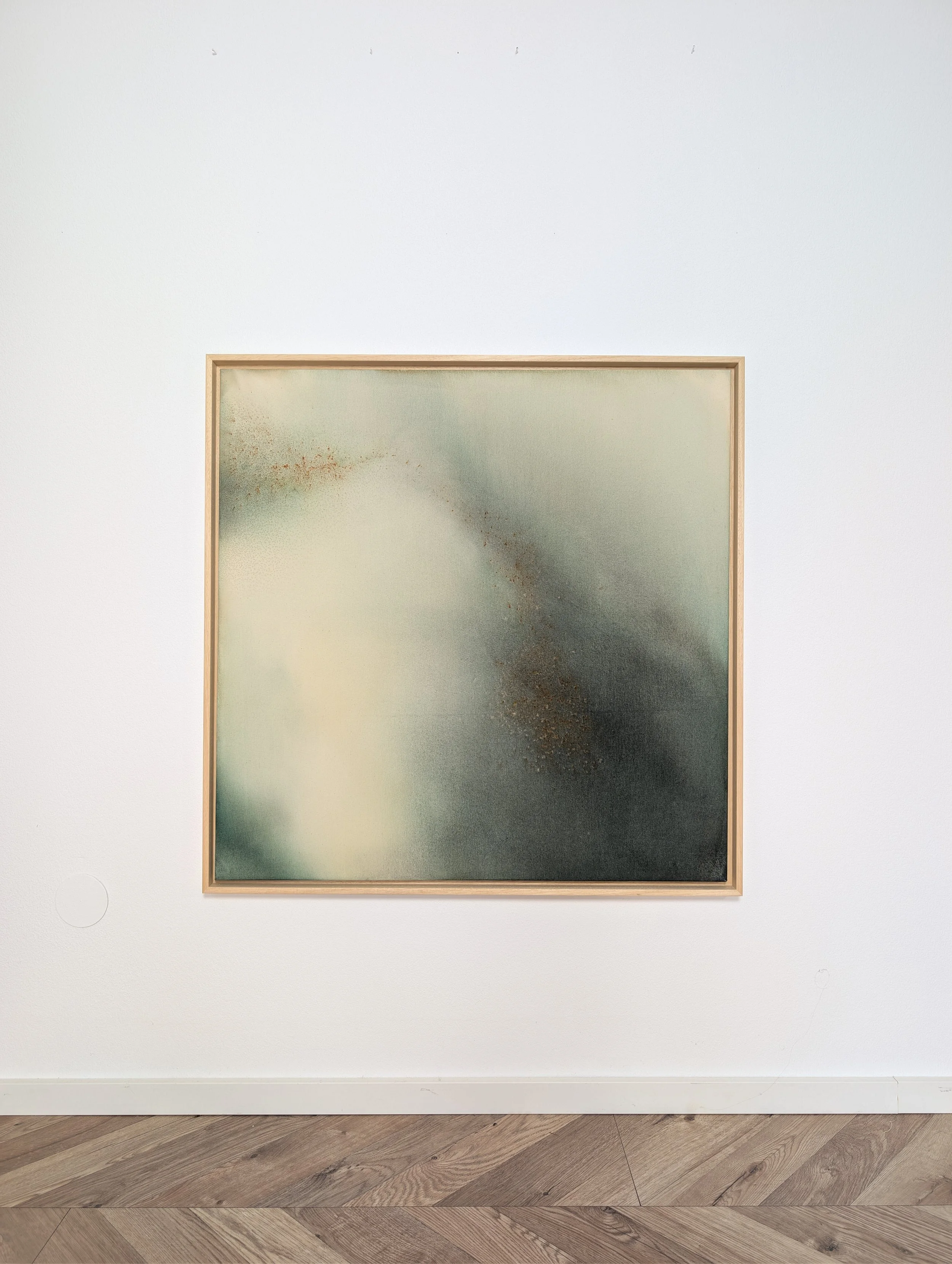 Kunstwerk in einem Rahmen vor weißer Wand, bestehend aus einer abstrakten, dezenten Farbmischung mit Texturen in Grau, Hellgrün und Senffarben, hängt in einem Ausstellungsraum.