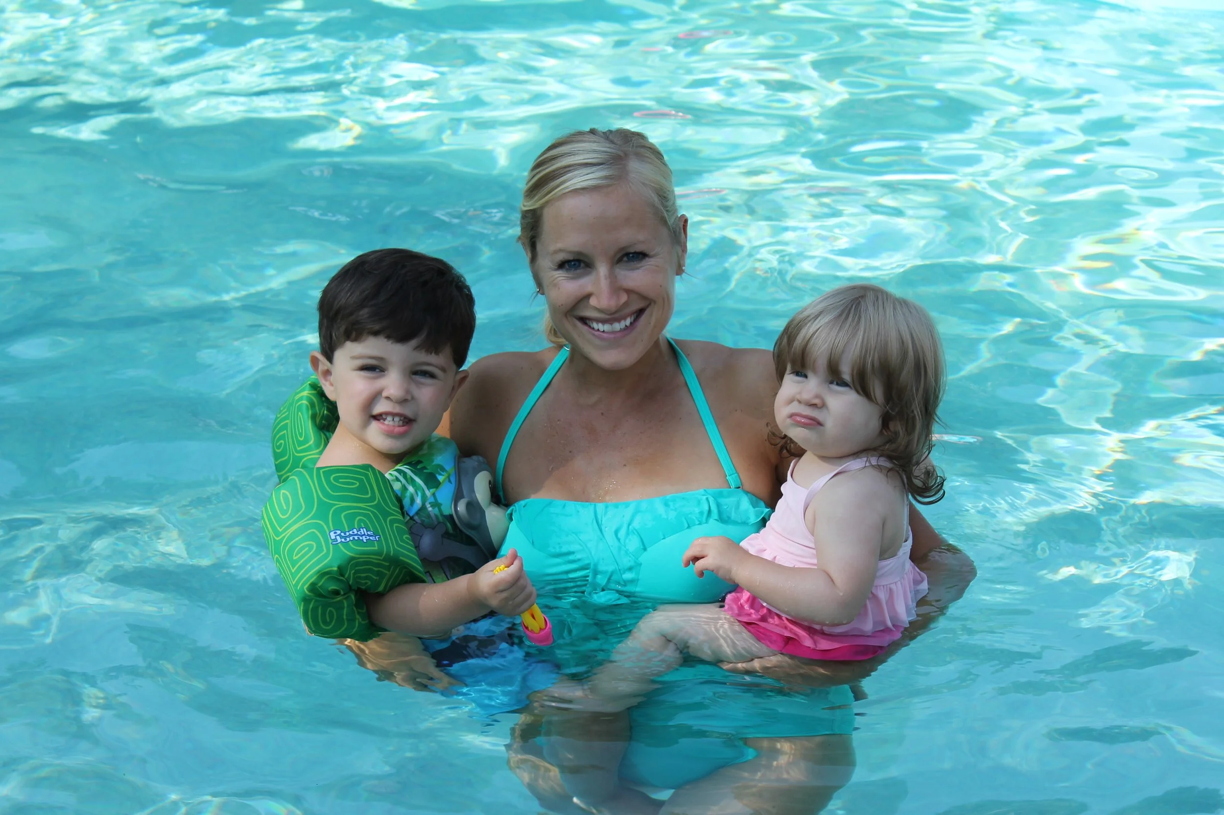 A woman in a blue swimsuit smiling and holding two young children in a swimming pool with clear blue water.