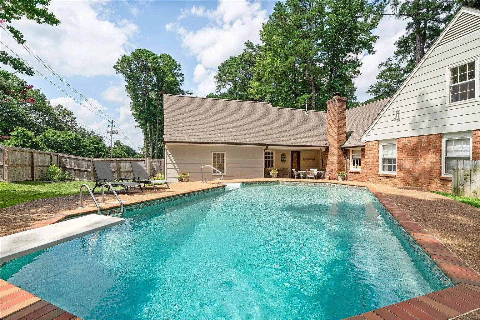 Residential backyard with a swimming pool, two lounge chairs, a patio table, and a house with white and brick exterior walls, trees, and a fence.