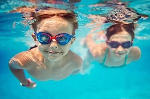 Two children swimming underwater in a pool, wearing goggles and smiling.
