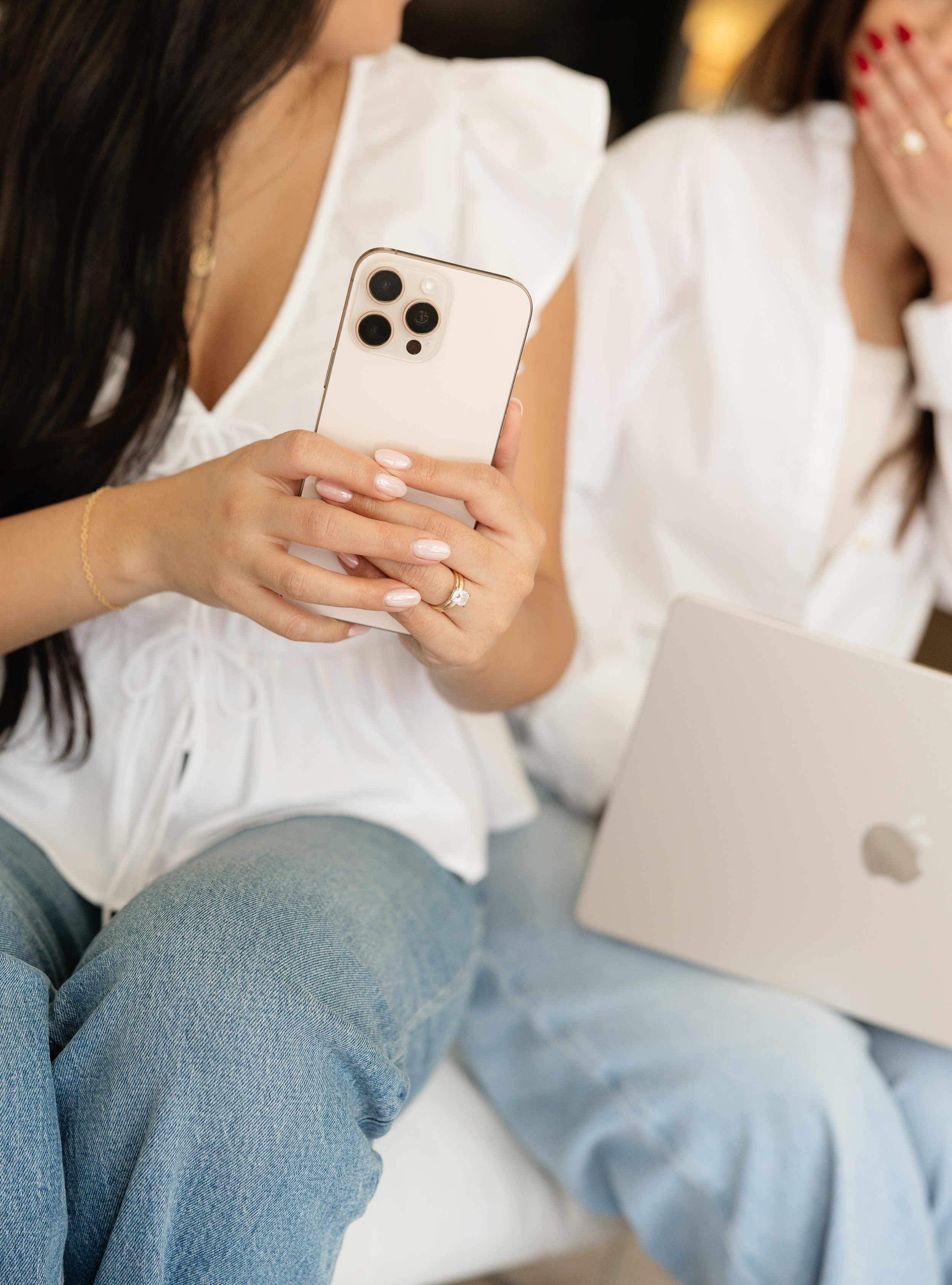 Close-up of a woman holding a white iPhone with three camera lenses, wearing a white top, gold jewelry, and light blue jeans, sitting with another woman in a white top who is covering her face with her hand, in an indoor setting.