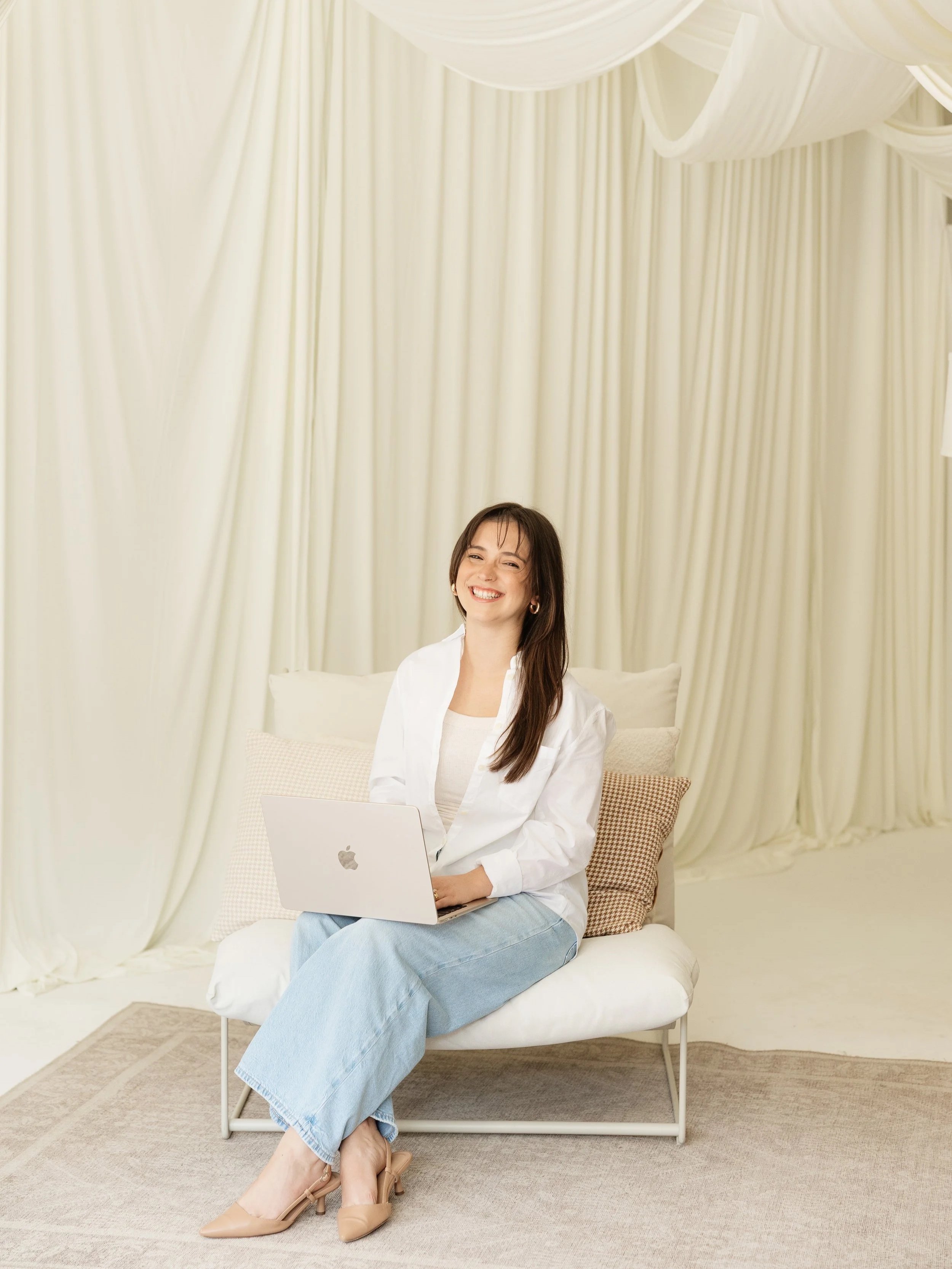 A woman sitting on a bed with cream-colored curtains in the background, working on a silver MacBook laptop, smiling with her legs crossed at the ankles, wearing a white top, blue jeans, and beige heels.