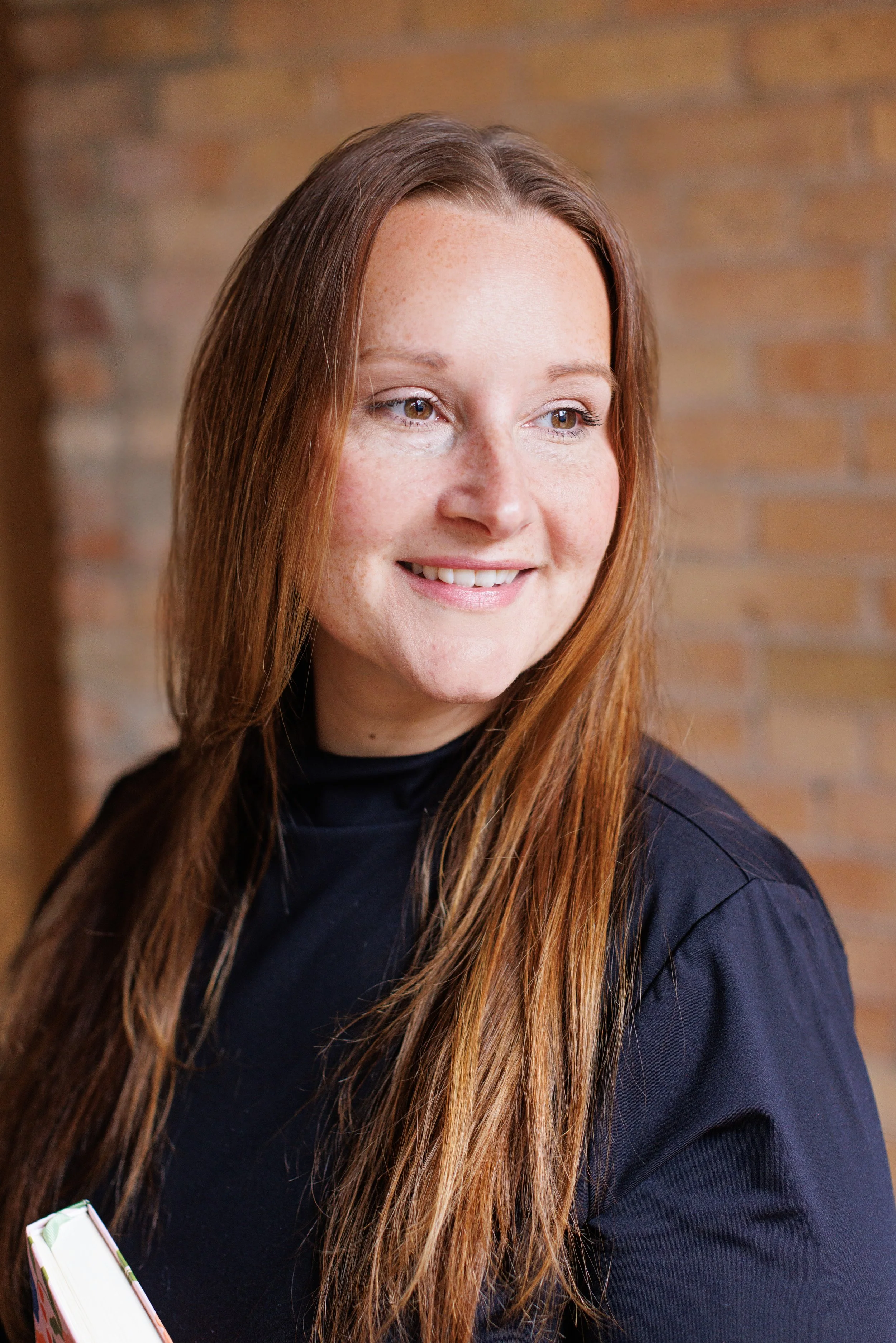 Close-up of a woman with long red hair, light skin, and freckles, smiling and looking to her left, positioned against a brick wall background.