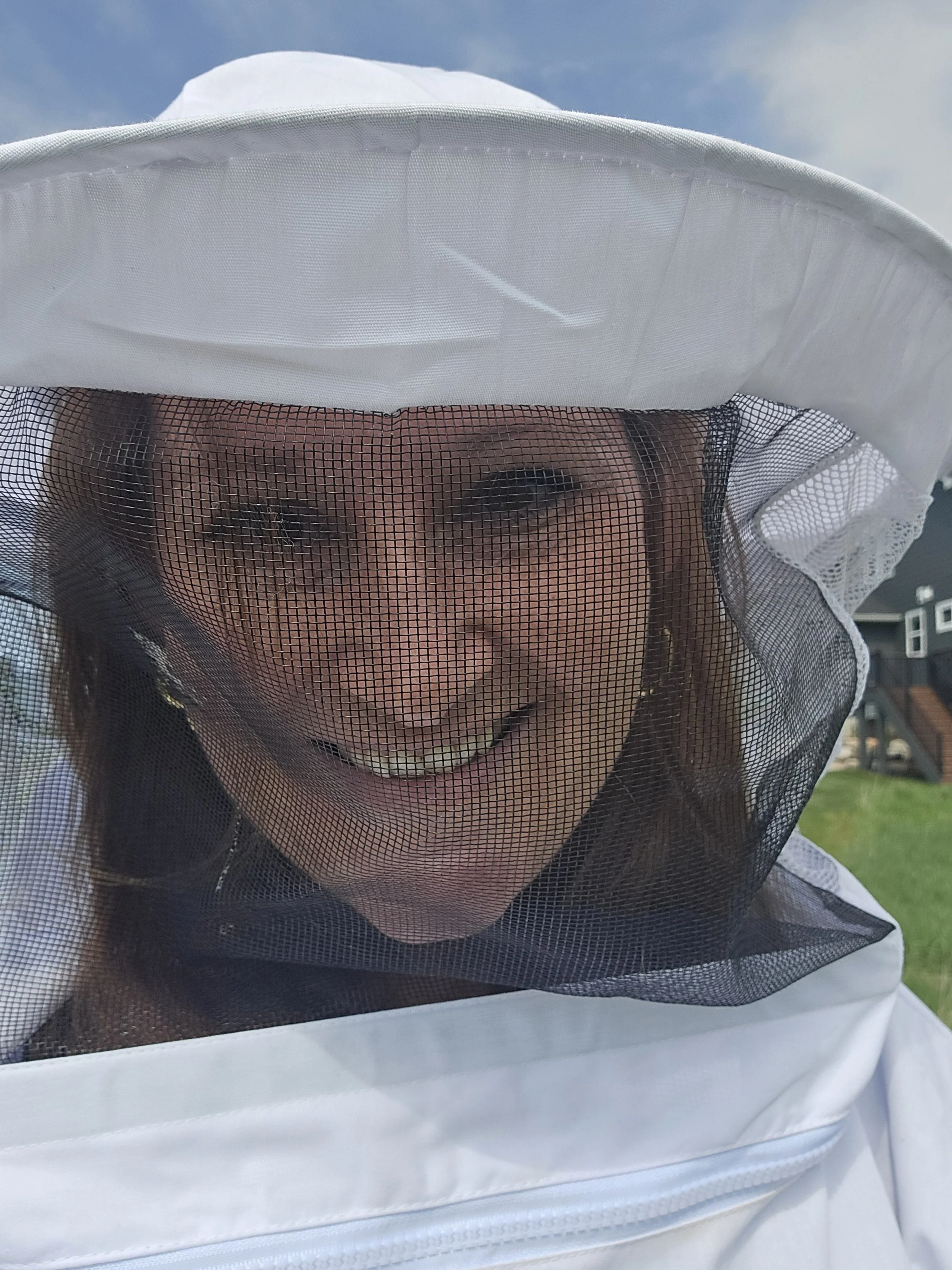 A person wearing a beekeeping suit with a mesh face shield, smiling outdoors under a blue sky.