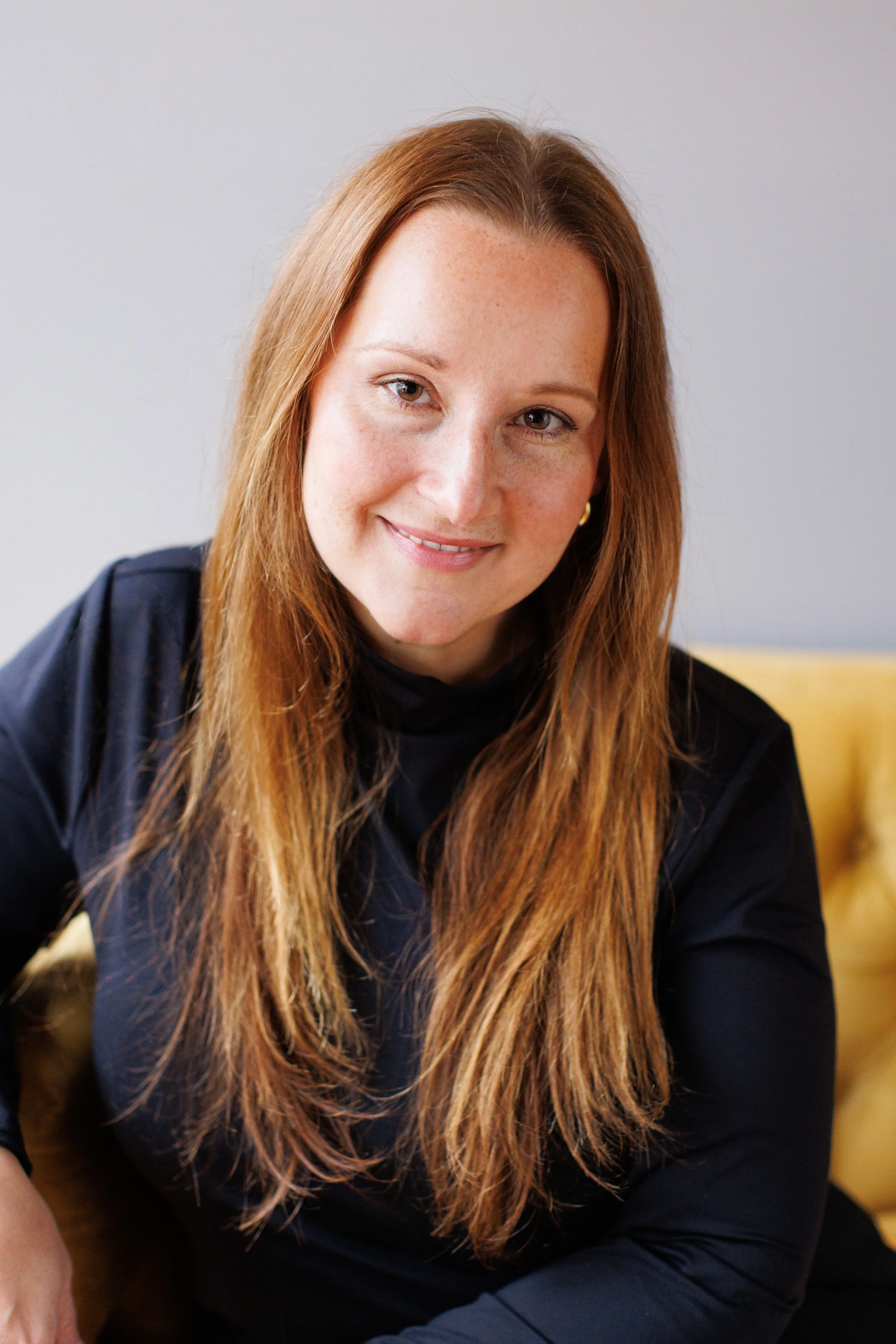 A woman with long red hair and light skin, wearing a dark top, smiling at the camera, seated indoors with a plain wall in the background.