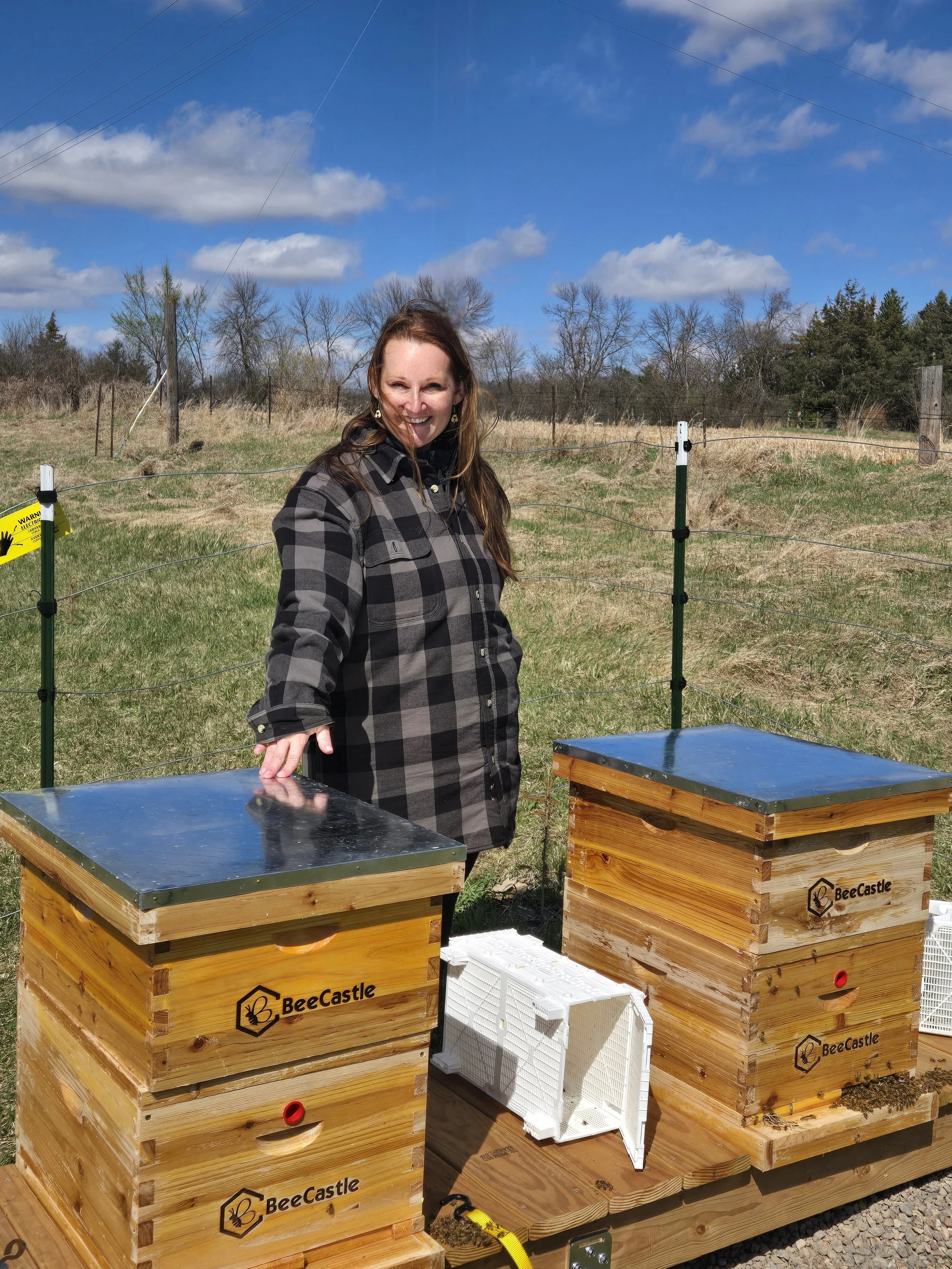 Woman standing next to two beehives labeled BeeCastle in an open field with trees and a blue sky in the background.