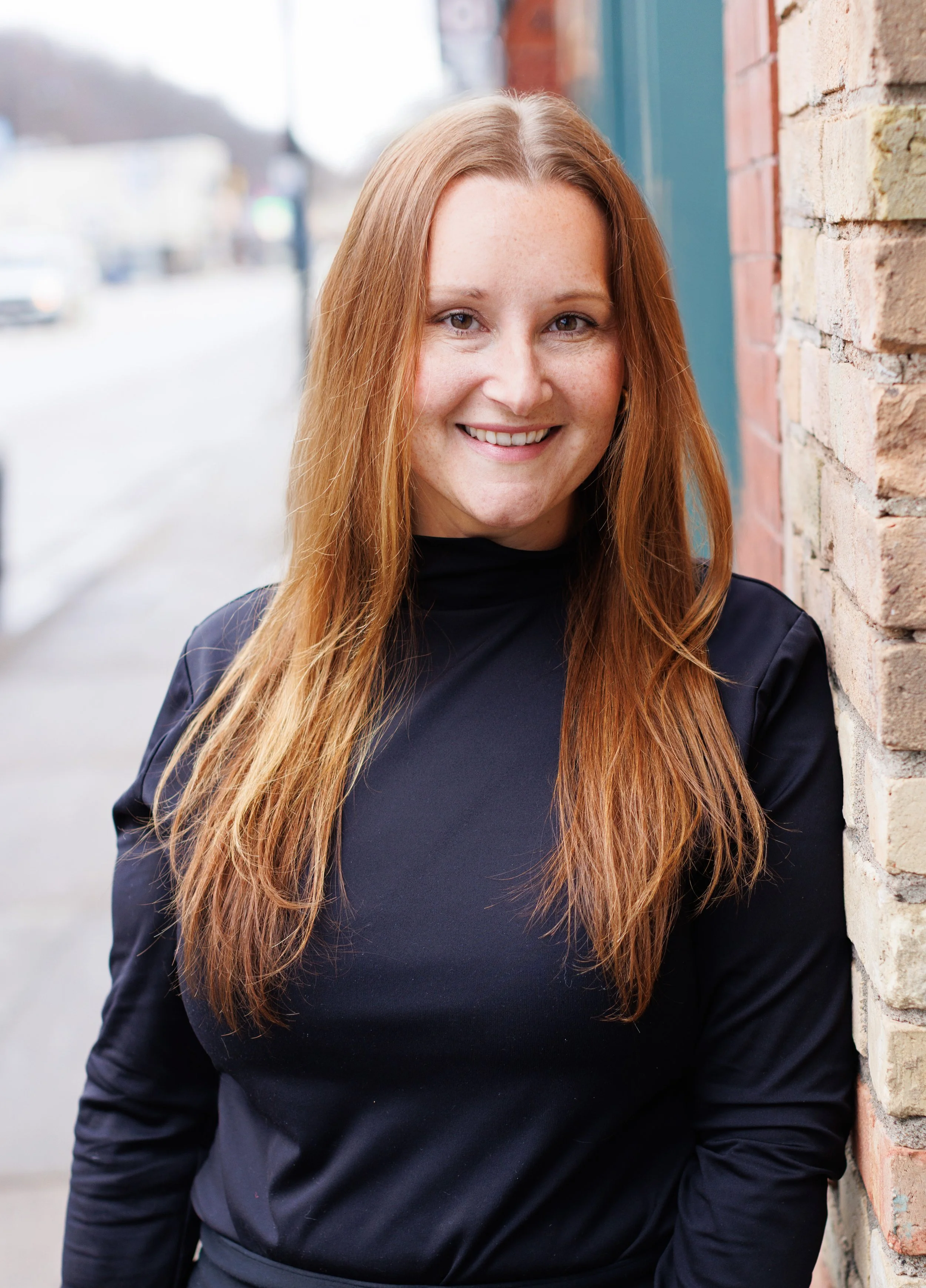 A woman with long red hair, smiling, standing outdoors next to a brick wall.