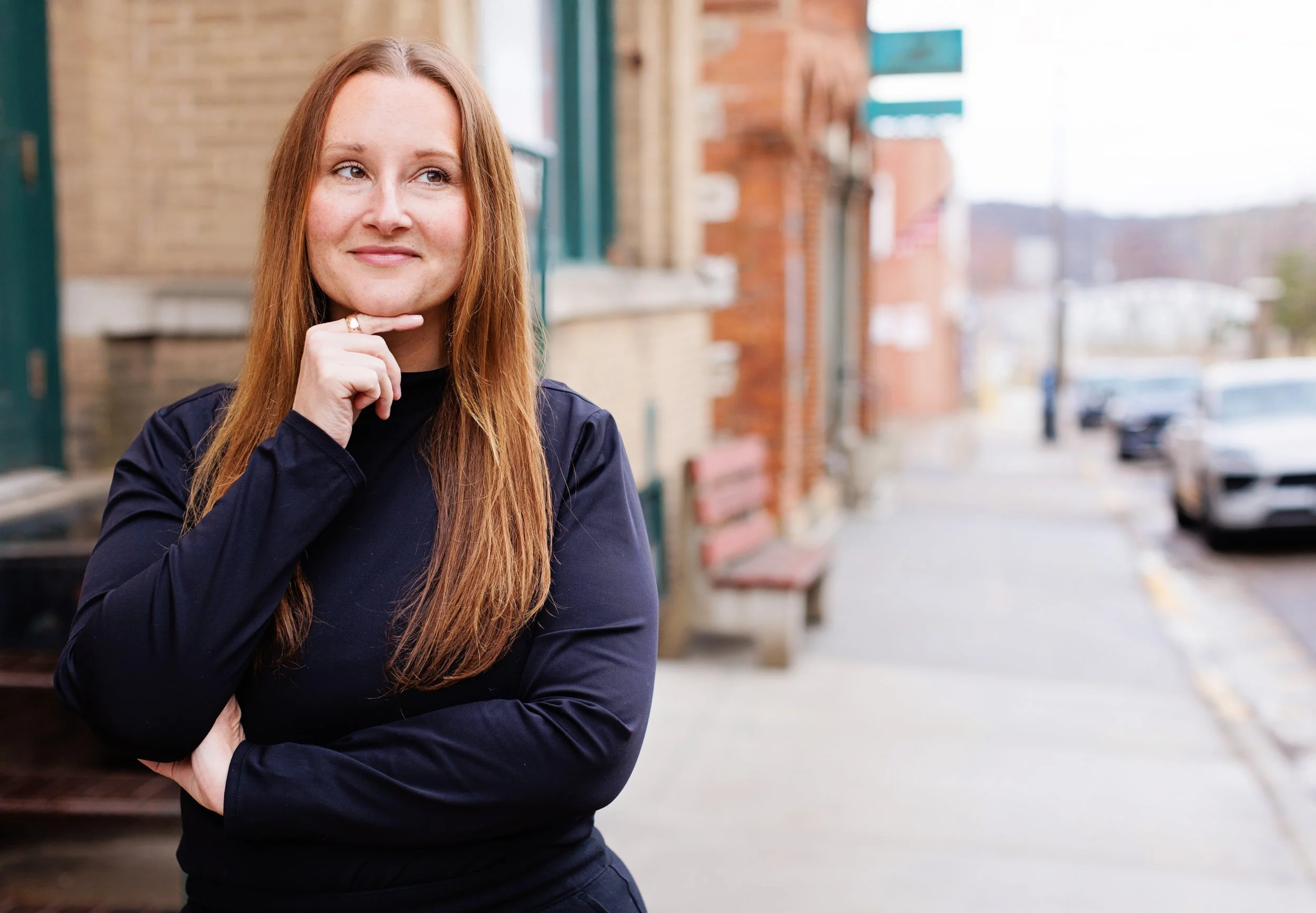 Red-haired woman in black jacket posing outdoors on a city sidewalk, with blurred buildings and parked cars in the background.