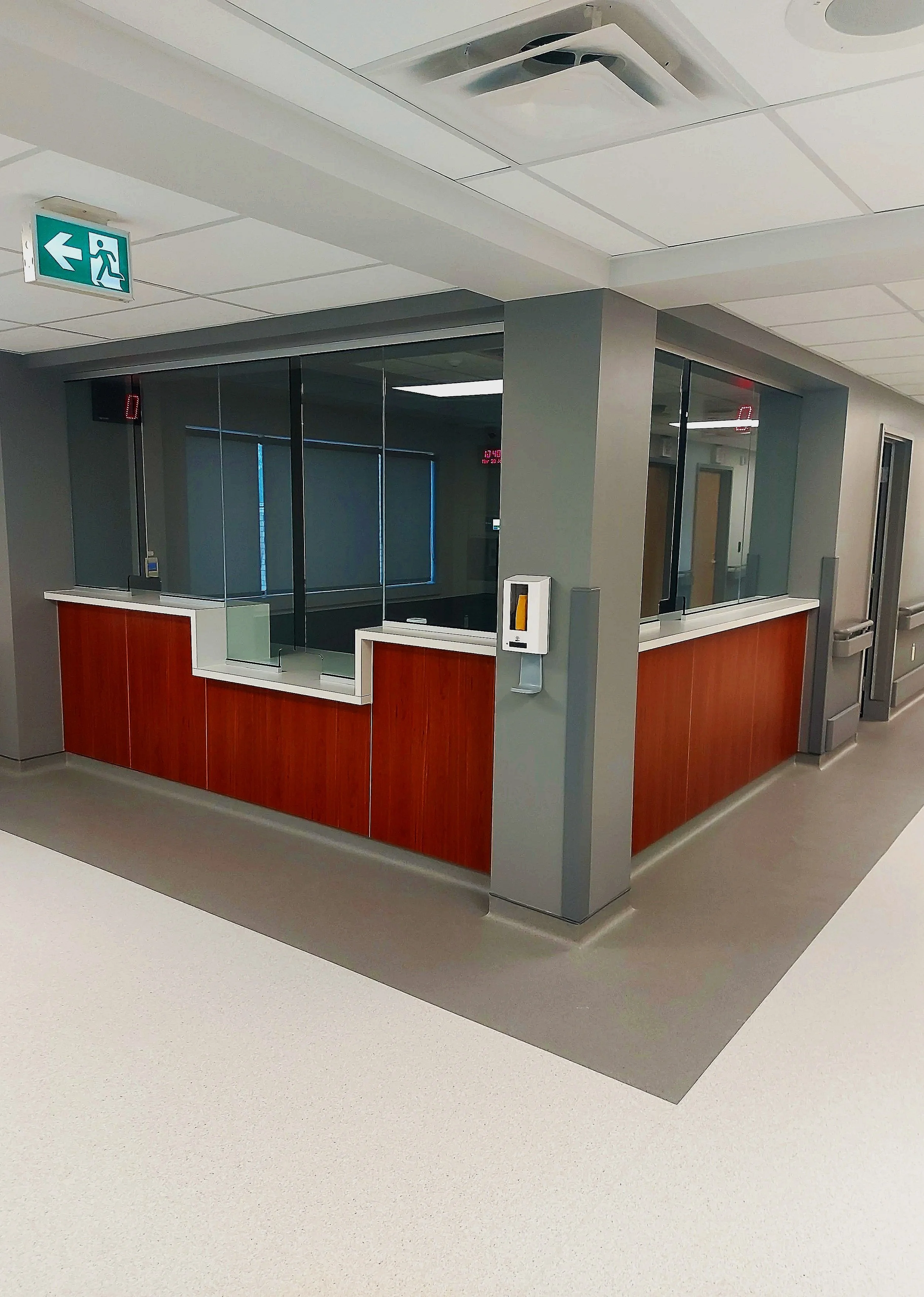 Empty hospital reception area with wooden counter, glass partitions, and an exit sign on the ceiling.