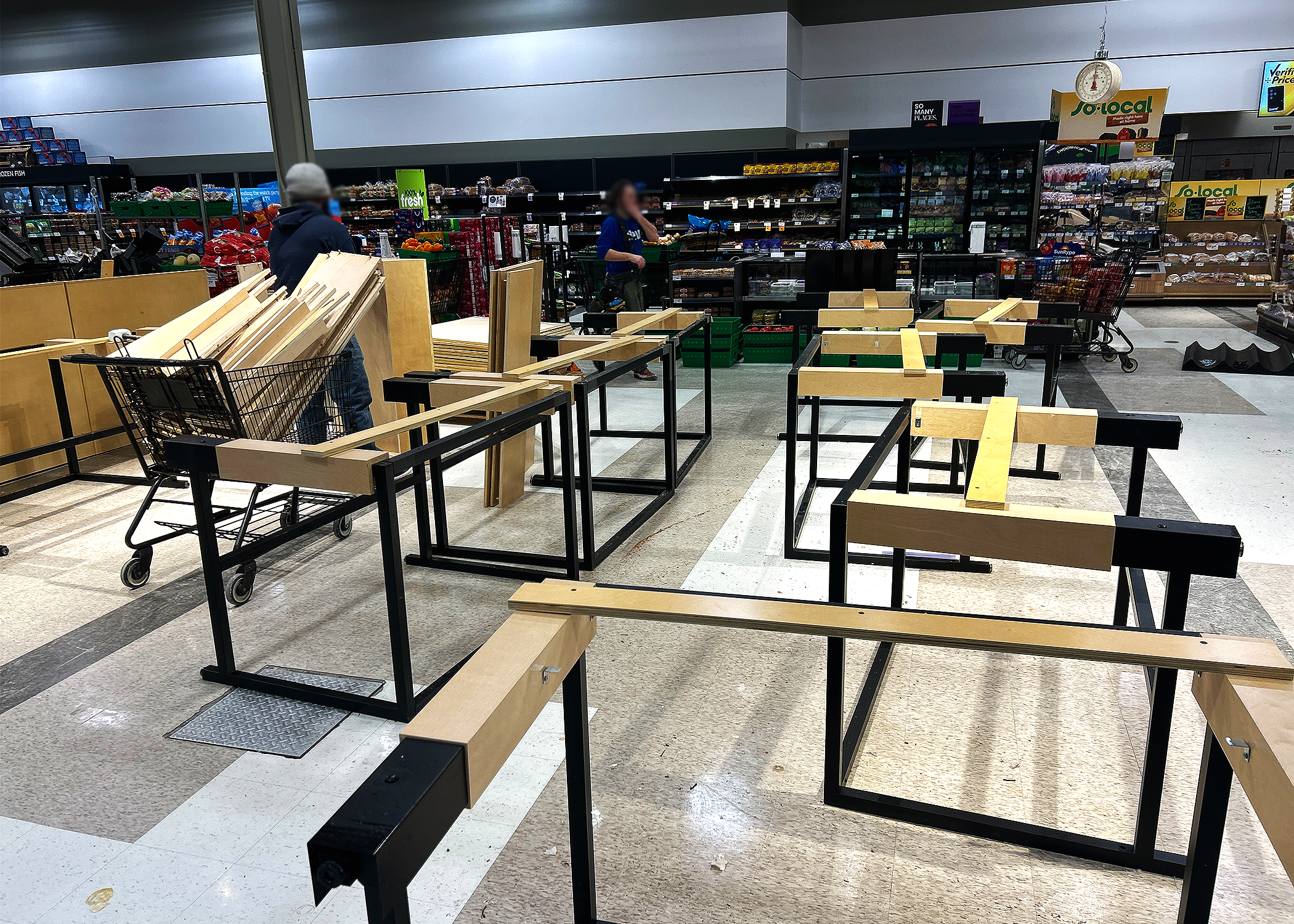 Inside a grocery store with empty shelving and wooden planks, shopping carts, and two people shopping.