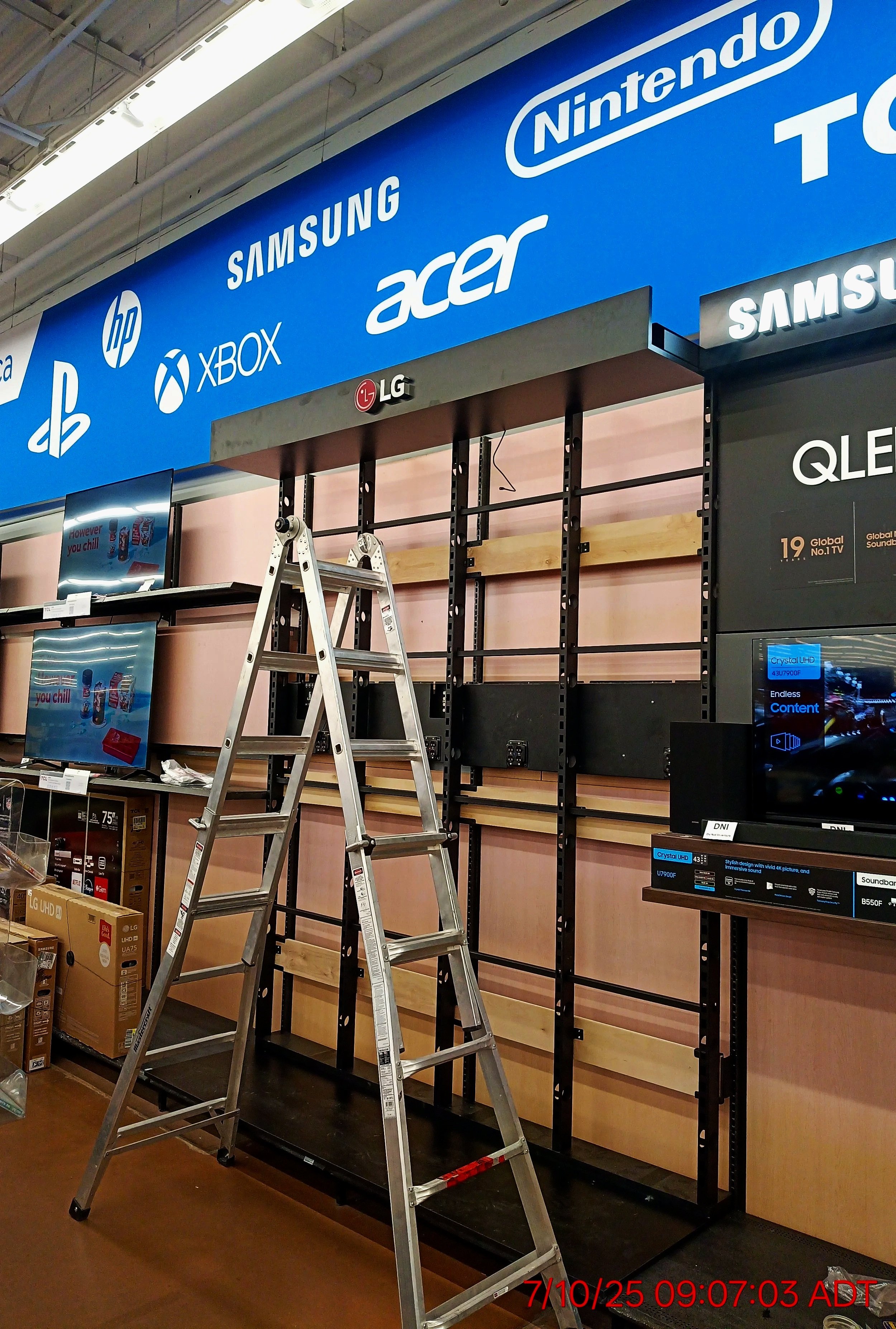 Empty retail space with black shelving units, a wooden background, a stepladder, and product boxes. Overhead sign displays logos of technology brands including HP, Samsung, Xbox, Acer, LG, and PlayStation. There are digital screens and packaging on the shelves.