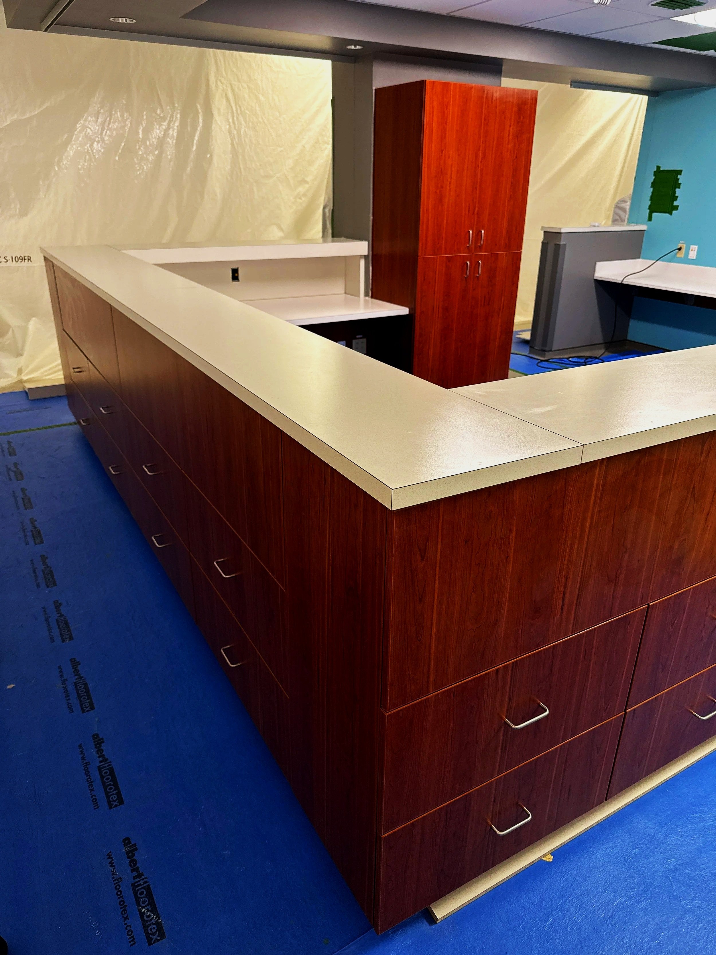 A U-shaped reception counter with wood grain finish and a white countertop, a tall wooden cabinet with doors, and a blue wall with a green wall decoration in an indoor setting.