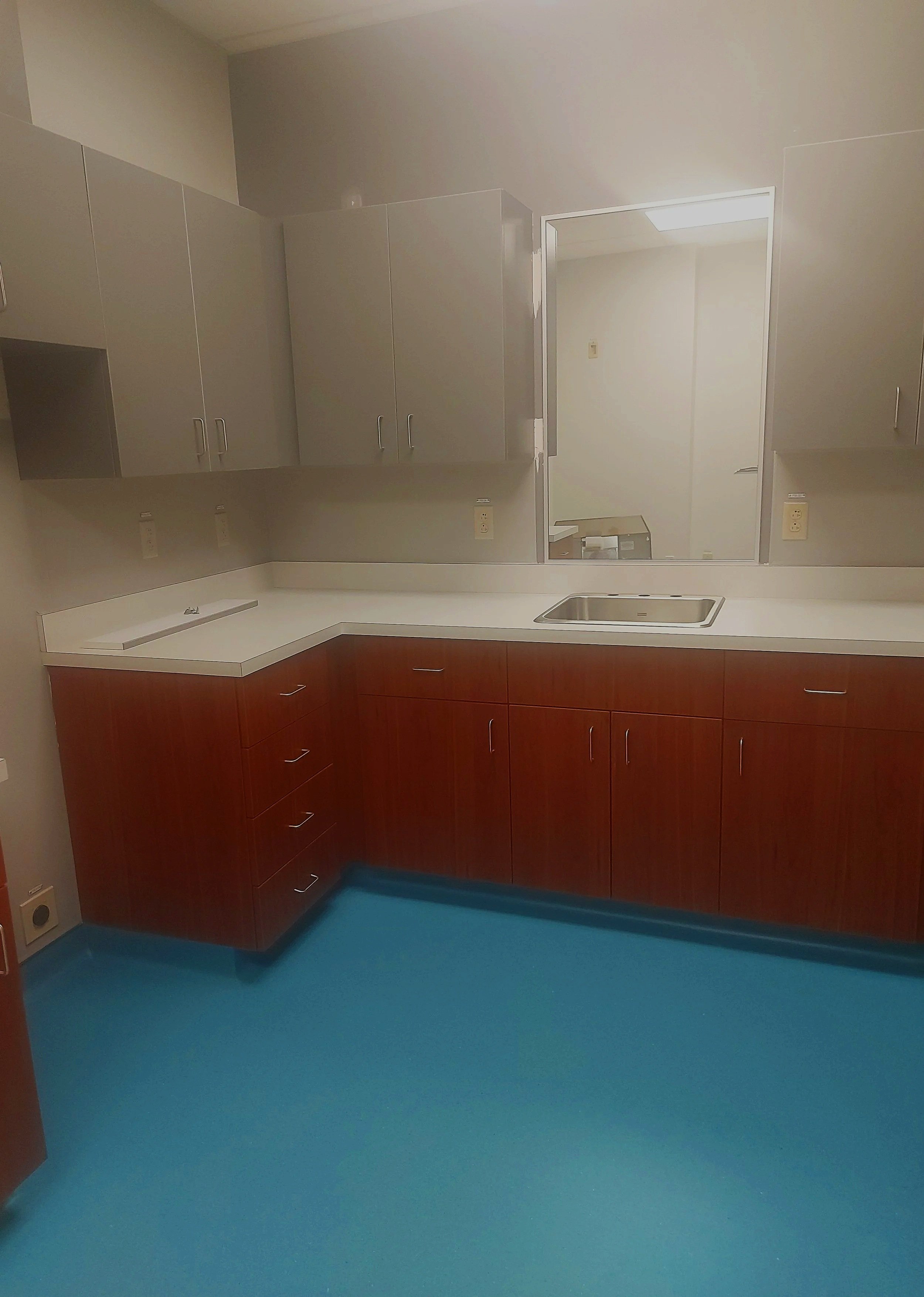 A kitchen with gray upper cabinets, a white countertop, a stainless steel sink, lower cabinets with a wood finish, and a blue floor.